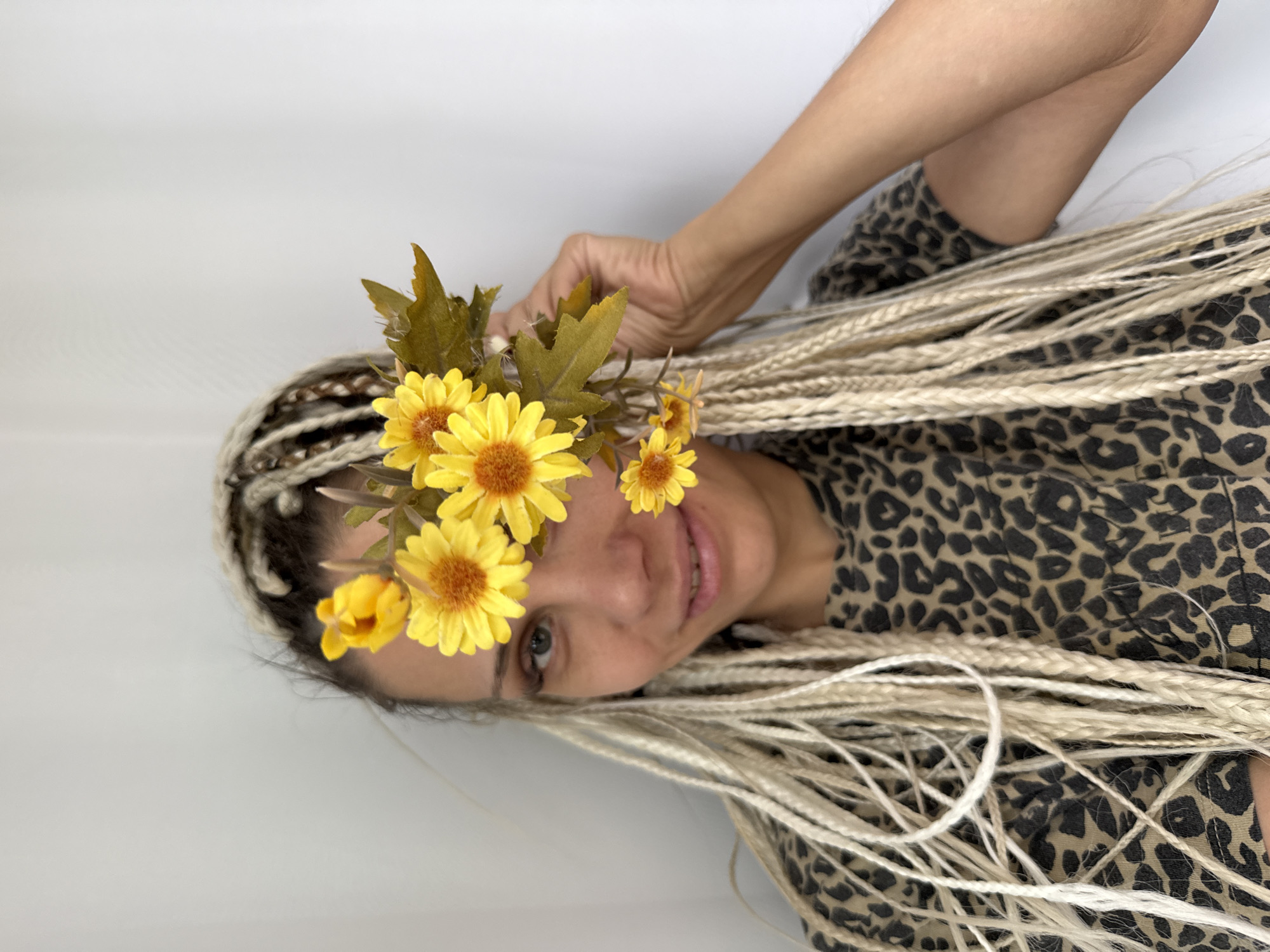 In Rusanovo, Russia, a young woman with blonde braided hair and a leopard-print shirt is shown. She is holding a bouquet of yellow flowers in front of her face, obscuring her left eye. The background is a plain white wall, suggesting an indoor setting, possibly a studio or a room in a residential building. The lighting is soft and diffused, casting no harsh shadows. Her expression is neutral, with a slight smile. The image is a close-up shot, focusing on the woman's face and upper body.