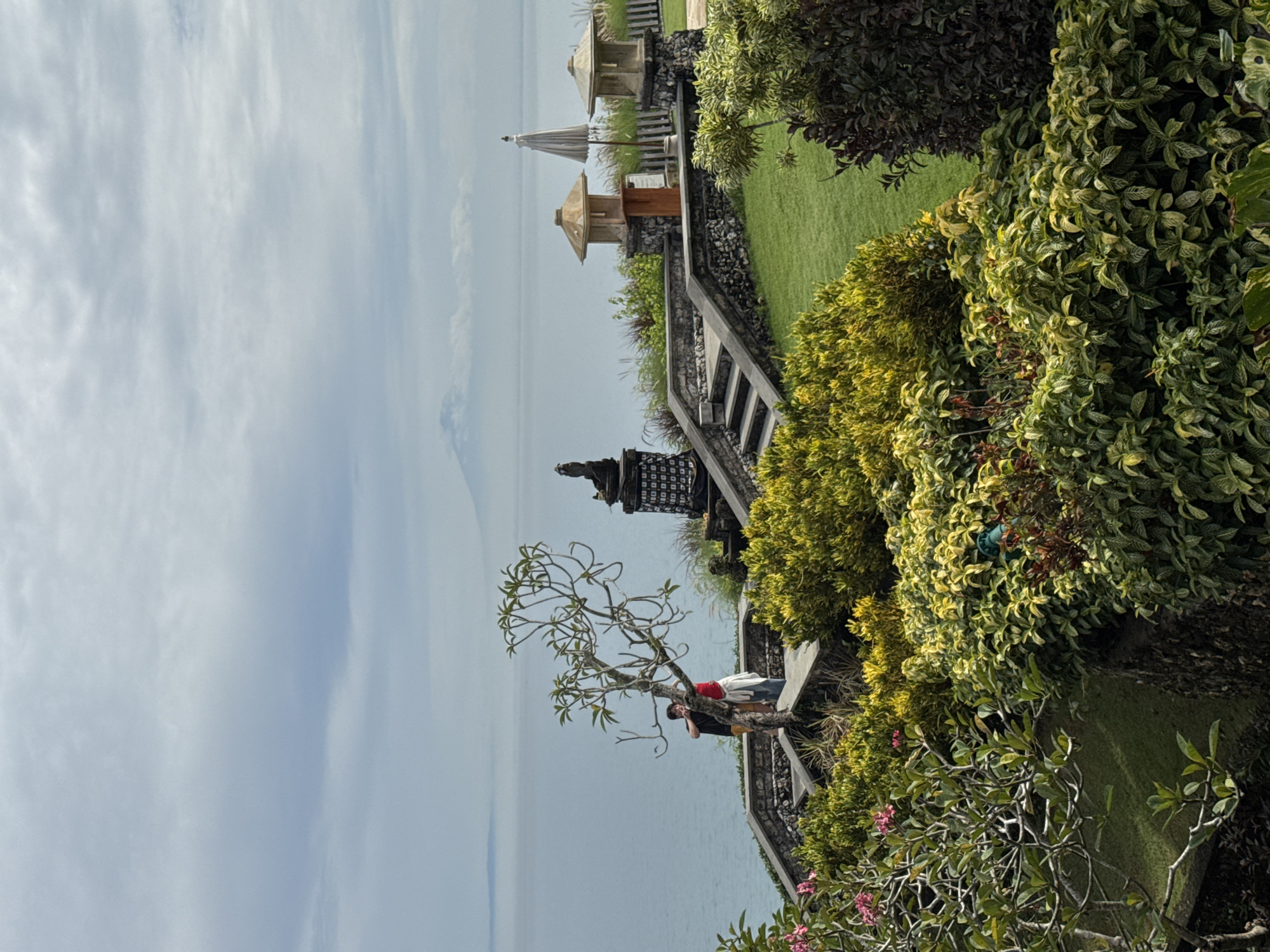 The image depicts a coastal scene in Kuta Selatan, Indonesia. A staircase descends towards the ocean, flanked by dense green and yellow foliage. A small tree with bare branches is visible near the railing, with two individuals standing behind it; one wearing a red shirt and the other a black shirt and yellow shorts. Further up the staircase are two small Balinese shrines or gazebos with traditional tiered roofs. In the background, the ocean stretches towards the horizon under a cloudy sky. A hazy landmass is faintly visible in the distance. A tall Balinese structure with intricate carvings is also visible near the staircase.