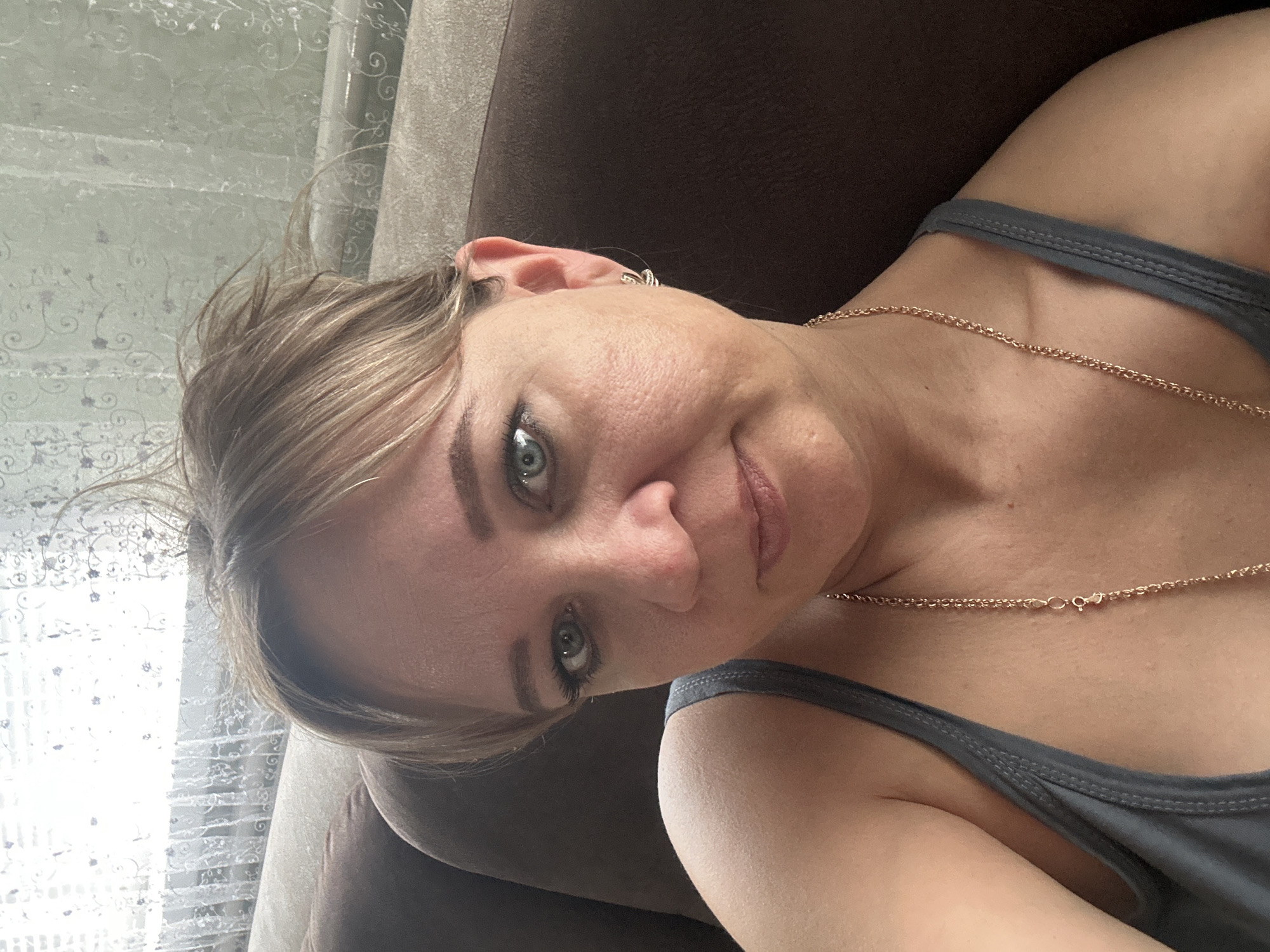 A middle-aged Caucasian woman with blonde hair is taking a selfie while sitting on a brown couch. She is wearing a grey tank top and a gold necklace. She is looking directly at the camera with a slight smile. The background shows a window with white lace curtains. The image appears to have been taken indoors in a residential setting.