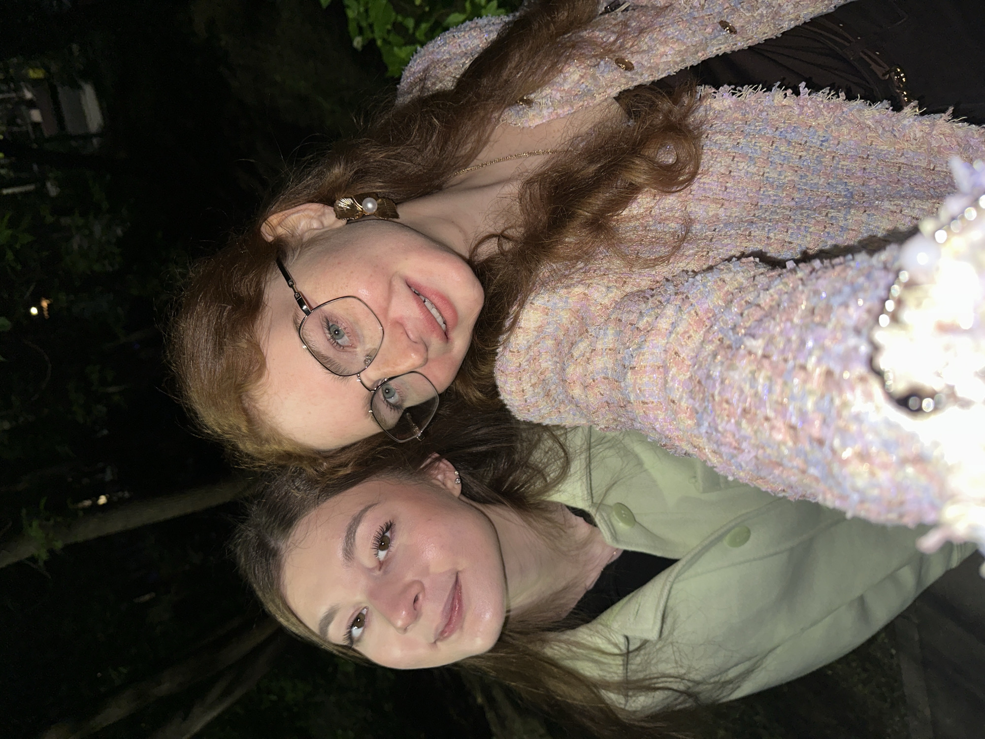 The image captures a nighttime outdoor selfie of two young adult females, presumably in the city of Kazan, Russia, given the contextual information provided.
The scene is dominated by the two individuals in the foreground, illuminated by a direct flash, which casts a bright light on their faces and clothing, while the background remains dark and out of focus, revealing silhouettes of trees and faint distant lights.
The female on the left, estimated to be in her late teens or early twenties, has long, light brown hair. She is wearing a light green, collared jacket over a dark top. Small, silver-toned stud earrings are visible in her earlobes. She looks directly at the camera with a gentle, closed-mouth smile.
The female on the right, also estimated to be in her late teens or early twenties, has long, wavy, reddish-brown hair. She wears square-framed eyeglasses, and her eyes appear light-colored. Prominent gold-toned, shell-shaped earlobe earrings, each featuring a white pearl, are visible. She is dressed in a textured, pastel-colored tweed-style jacket with small gold buttons, worn over a dark garment, and a thin gold-toned necklace. She smiles broadly, looking directly at the camera. Her skin exhibits freckles.
Both subjects are positioned closely together, facing the camera, indicating a self-portrait. A bright, out-of-focus object, possibly a decorative element or part of the camera itself, is present in the lower center foreground. The overall environment is an undifferentiated outdoor setting, likely a park or garden, at night.
