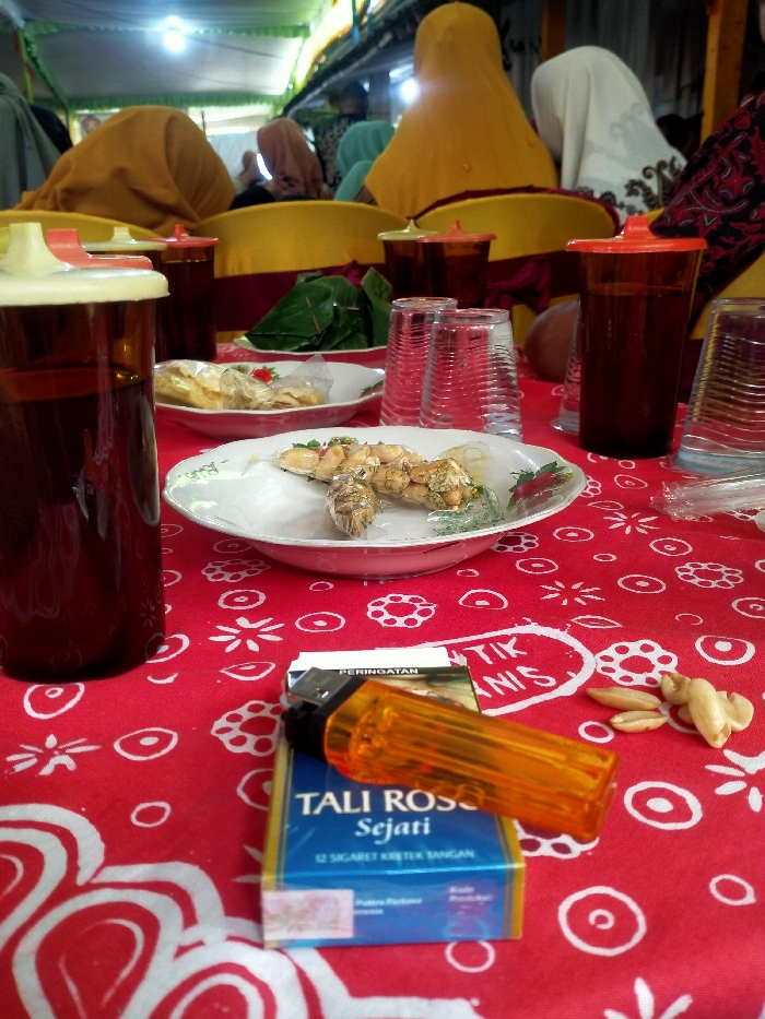 A red tablecloth with white patterns covers a table set with food, drinks, and cigarettes. In the foreground, a pack of "TALI ROSSO Sejati" cigarettes and an orange lighter sit next to a scattering of peanuts. Dishes hold various foods, and drinks are in brown plastic pitchers and clear glasses. In the background, people wearing hijabs are seated at the table. The scene appears to be a social gathering.