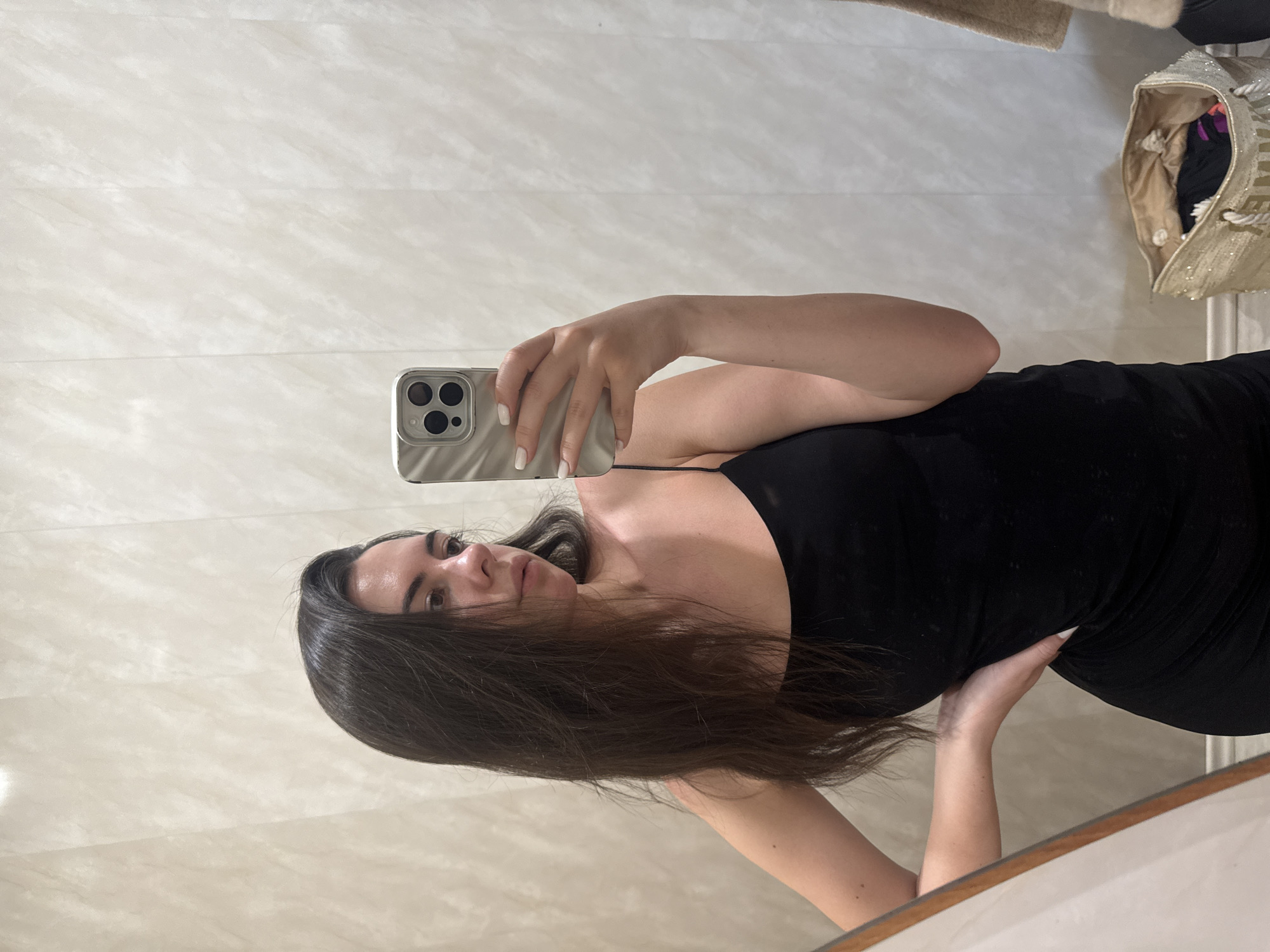 A young woman with dark brown hair is standing in what appears to be a fitting room, taking a mirror selfie with her phone. She is wearing a black spaghetti strap dress and has her left hand on her hip. Her right hand holds a silver smartphone, reflecting in the mirror. The background consists of a textured, light-colored wall and a glimpse of a large, open tote bag filled with items on the floor to the right.