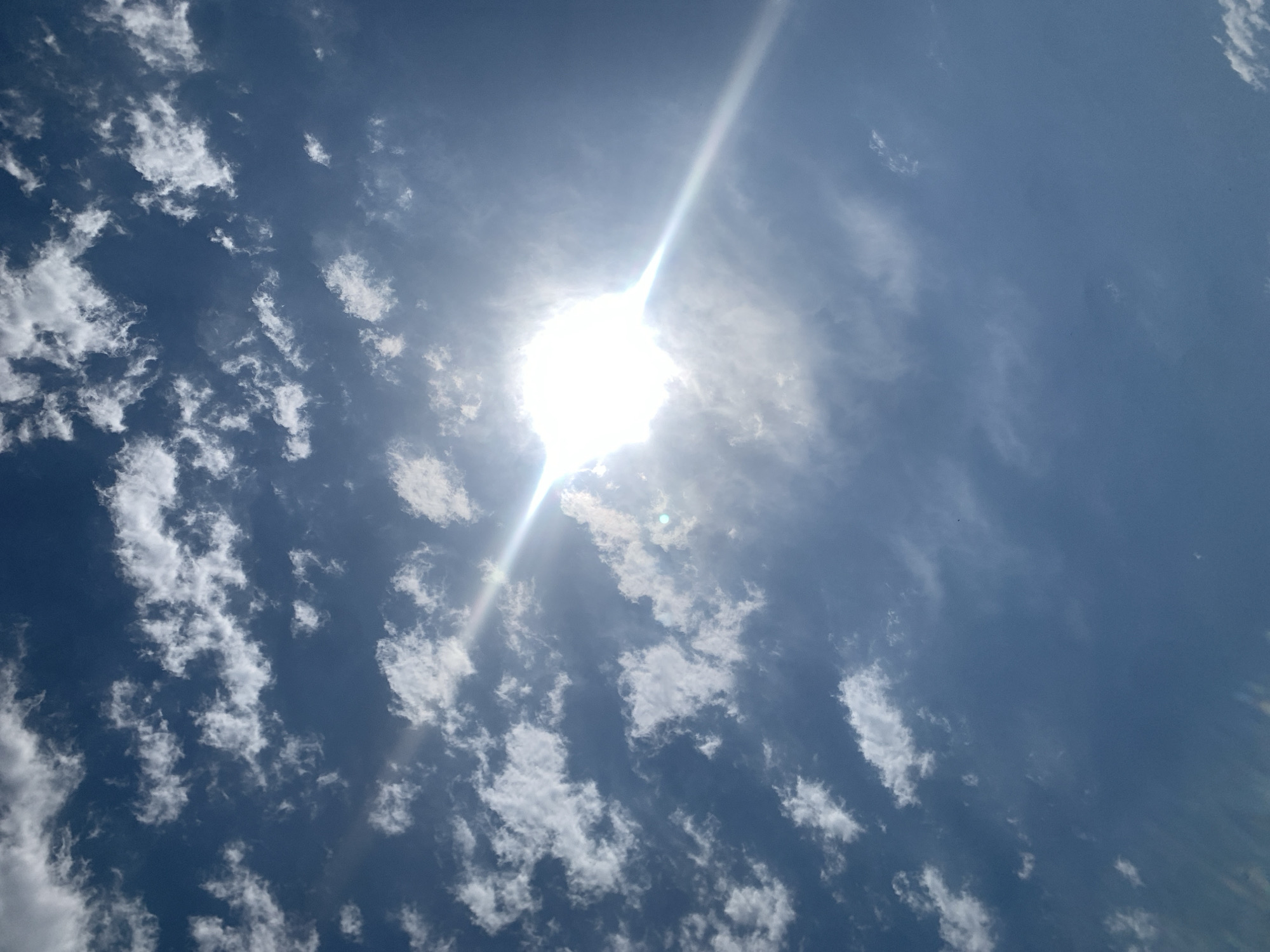 The image shows a bright sun shining through scattered white clouds against a deep blue sky. A sunbeam extends diagonally from the bright sun towards the bottom right of the frame. The scene is outdoors, with no visible structures or people. The location is identified as Khazar Raion, Azerbaijan, suggesting an open sky environment within that region.