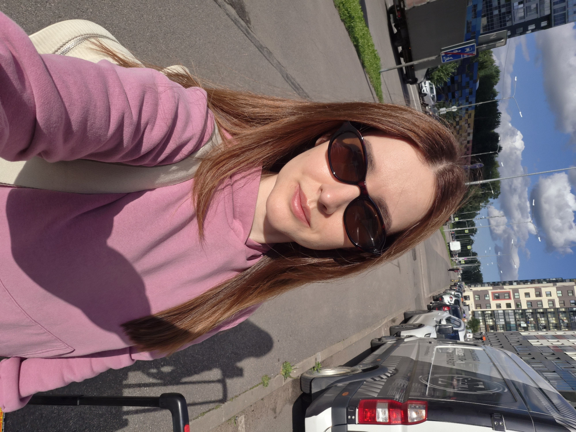 A young woman with long, auburn hair and sunglasses takes a selfie outdoors on a sunny day in Kudrovo, Russia. She is wearing a pink hooded sweatshirt and a light-colored bag is slung over her shoulder. To her right, a dark-colored suitcase with a red handle is visible. The background shows a street with several parked vehicles, including a white van with graphics. In the distance, a tall, modern apartment building dominates the skyline, and a truck and trees line the street. The scene appears to be a sidewalk adjacent to a road.