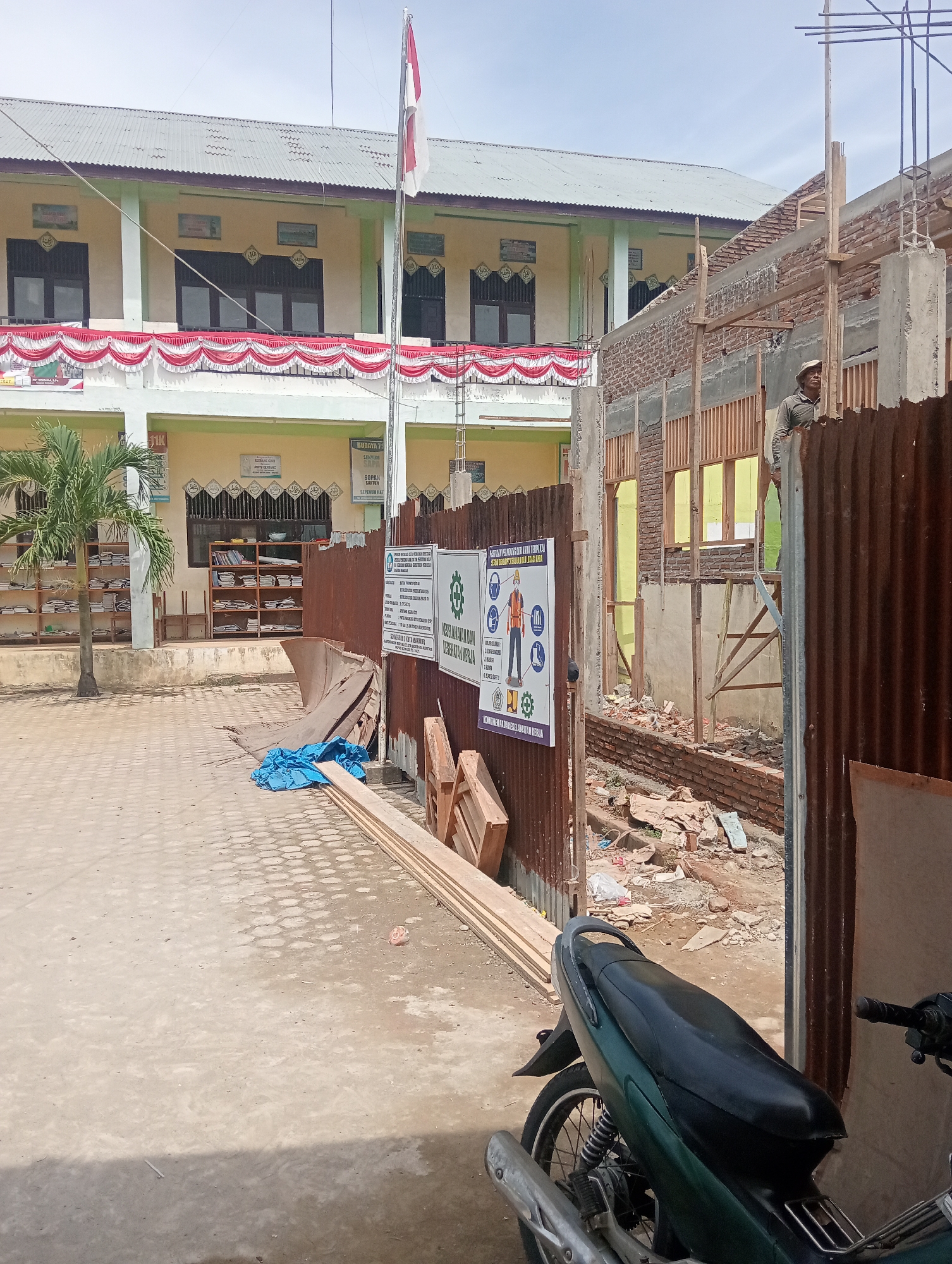 The image depicts a construction site adjacent to a school building in Keudekrueng, Indonesia. A two-story school building, painted in yellow and green, is visible on the left. Red and white decorative banners are draped along the second-story balcony. In front of the school, a small palm tree stands next to rows of shelves filled with books, suggesting a library or classroom area.
The right side of the image shows a building under construction. Exposed brickwork, wooden scaffolding, and concrete pillars are evident. A man, wearing a hat and a grey long-sleeved shirt, stands on the construction site, observing. Corrugated metal sheets form a temporary fence around the construction area. Safety posters are attached to the fence, one displaying safety regulations with illustrated figures.
In the foreground, a green motorcycle is parked on a paved walkway. Piles of lumber and a blue tarp are also visible on the ground, indicating ongoing construction work and material storage. The overall environment suggests a sunny day with a clear sky. Debris and construction materials litter the ground within the construction zone, highlighting the active nature of the site.