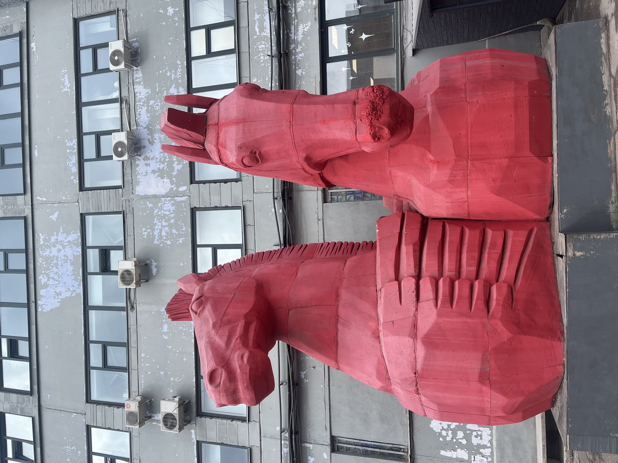 Two large, red, geometric horse head sculptures are positioned next to a gray building with rows of windows and multiple air conditioning units. The building appears to be weathered with peeling paint. The sculptures are constructed from angular pieces and have a rough, textured surface. The location is Krasnoyarsk, Russia.