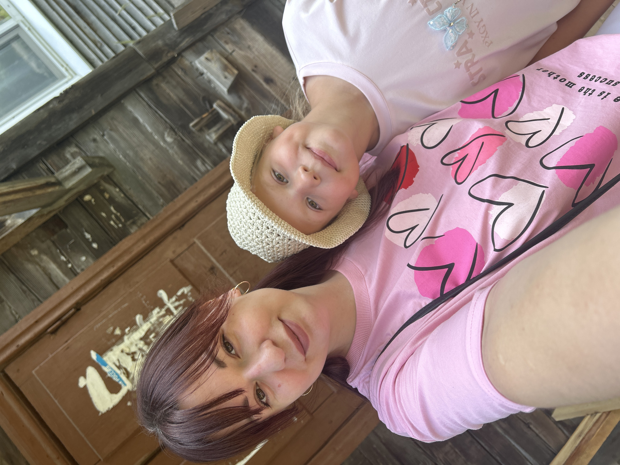 A selfie shows two people, an adult and a child, standing in front of a weathered wooden building. The adult, a woman with dark hair and bangs, is on the left, wearing a pink t-shirt with a heart design. She is looking at the camera. A young girl, with blonde hair and a straw hat, is leaning her head on the woman's shoulder, looking towards the camera with a smile. She is wearing a light pink t-shirt. The wooden building behind them has a door and what appears to be a window. The building's wood is aged and shows signs of wear. The overall setting suggests an outdoor location, possibly in a rural or older part of the city of Krasnovishersk, Russia.