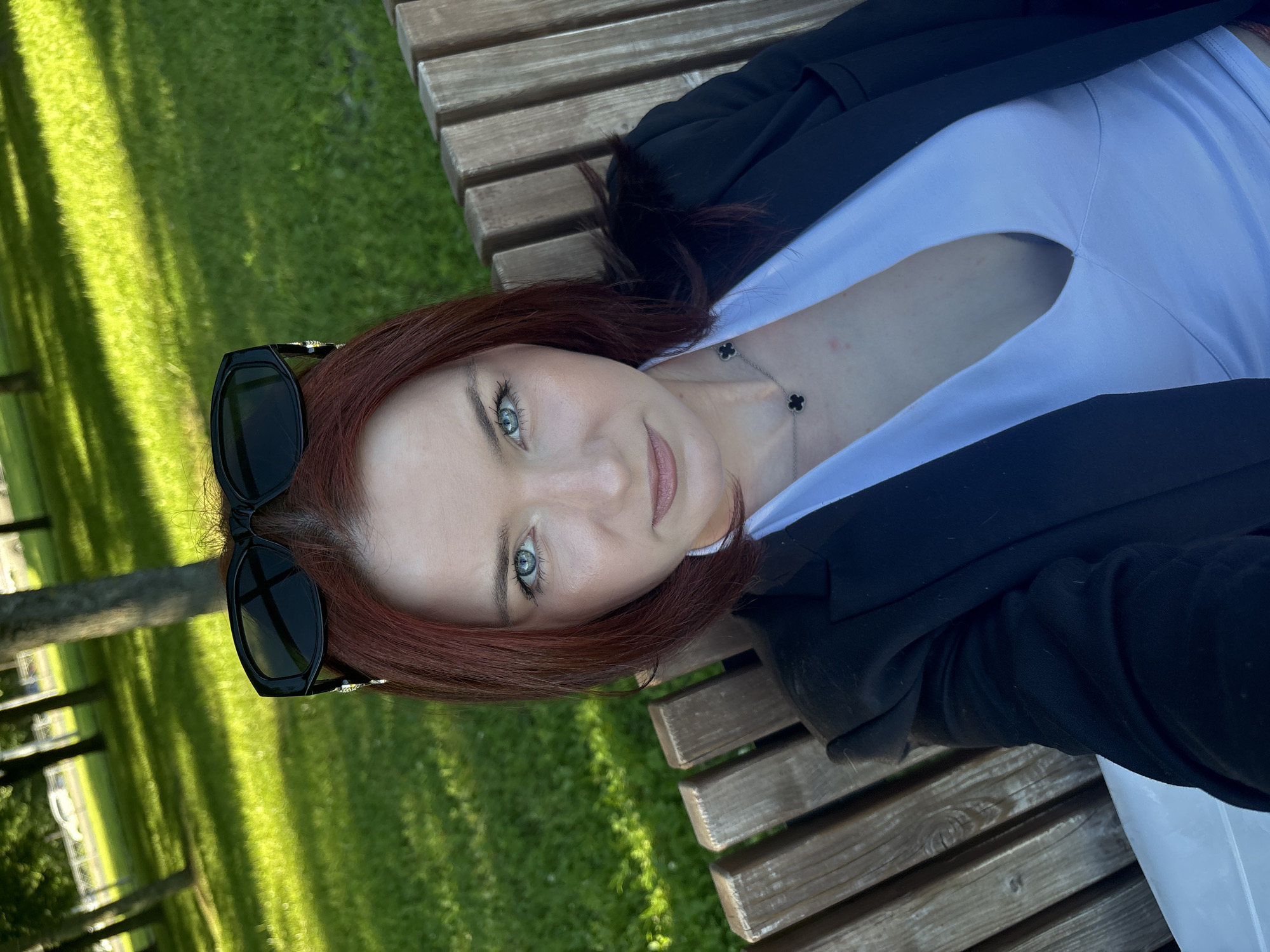 A woman with red hair is sitting on a park bench in Sinyavino, Russia. She is wearing black sunglasses on top of her head, a black blazer, and a white V-neck top. She is looking at the camera with her blue eyes. The background is a grassy park with trees and sunlight filtering through them. The bench is made of wooden slats.