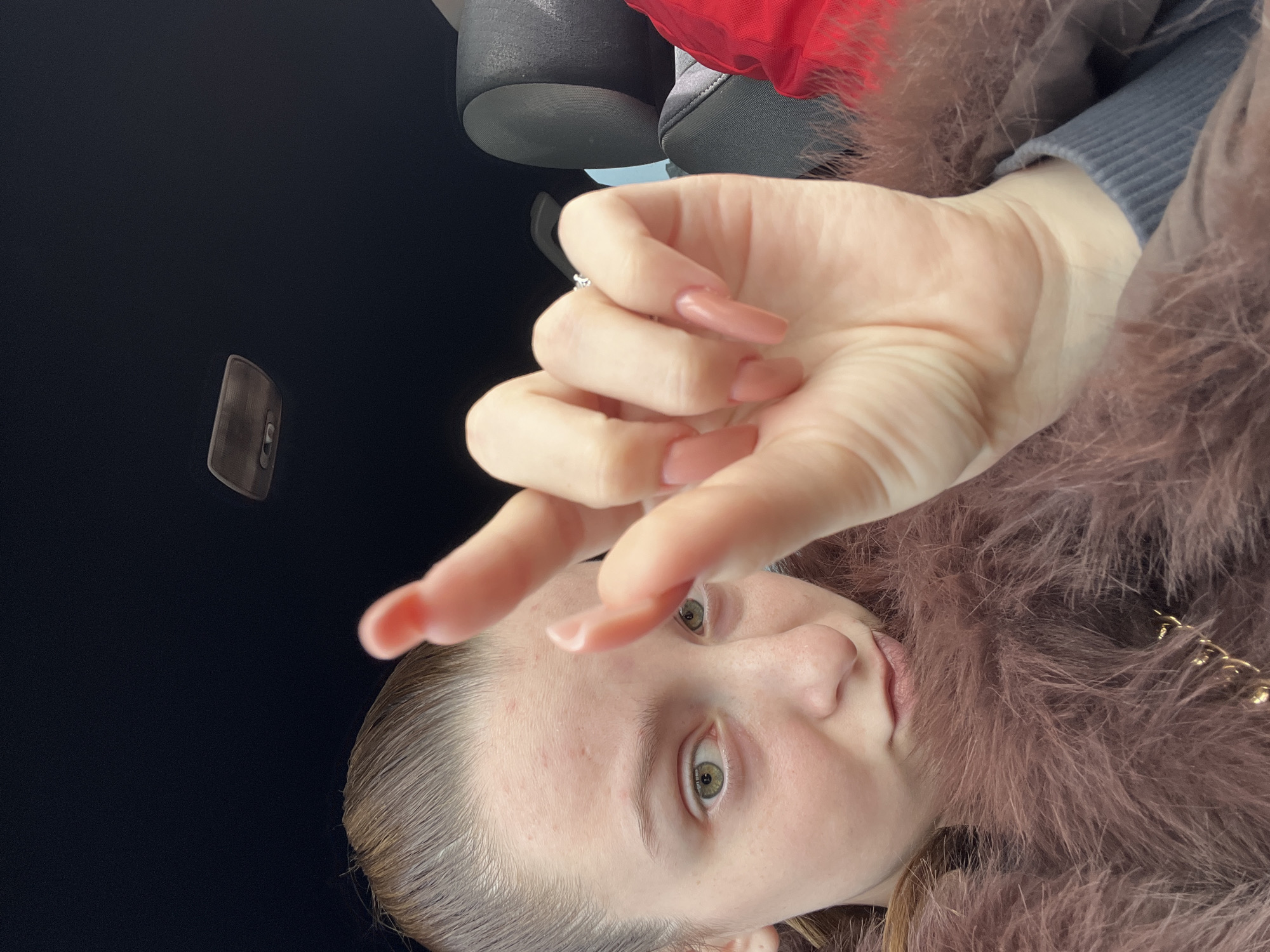 A selfie taken inside a car, under an overcast sky. The subject, a young woman, is looking directly at the camera, holding her hand up in front of her face. She has pale skin, blonde hair slicked back, and green eyes. She is wearing a furry mauve colored jacket. Her fingernails are long and painted a nude pink. There is a grey car seat visible in the upper portion of the frame. The car's interior light panel is affixed to the dark ceiling. The location is assumed to be in Varna, Bulgaria. No specific details confirm the precise location within the city.
