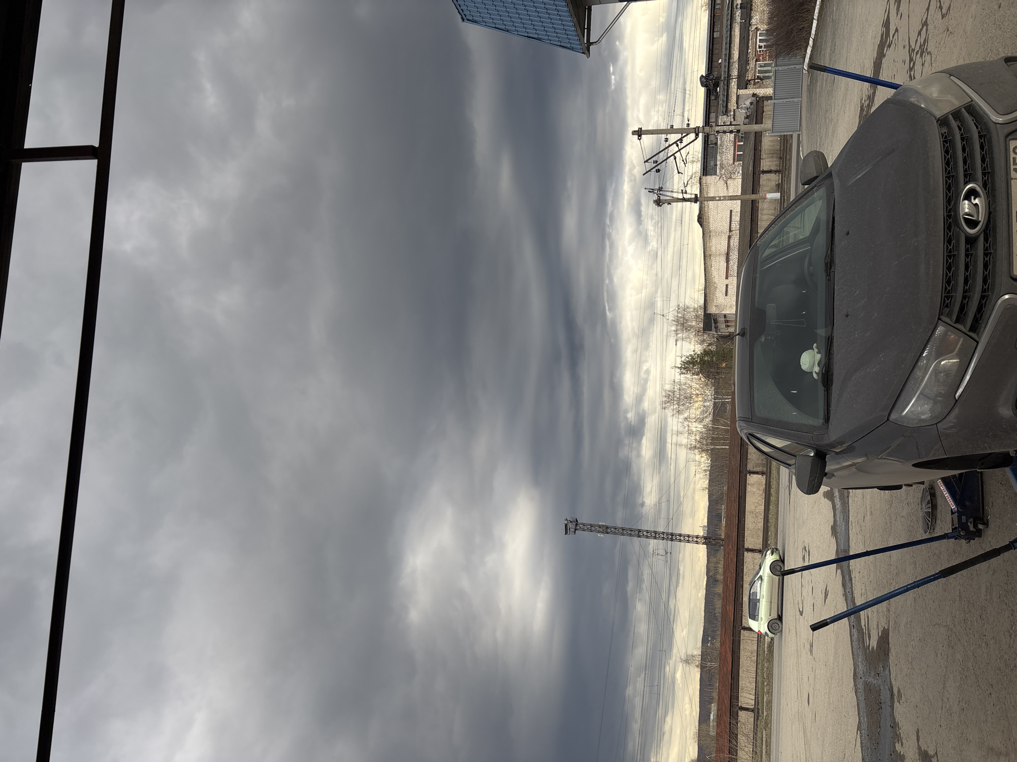 This outdoor image captures an industrial or semi-urban scene under a heavily overcast sky. A dark-colored car, appearing to be a Lada XRAY based on the visible grille emblem, is prominent in the foreground on the right. It is elevated, with a blue hydraulic jack placed underneath and two long blue support rods extending to the ground, suggesting vehicle maintenance or a tire change is in progress. The ground is a worn asphalt or concrete surface.
In the mid-ground, a smaller, light-colored hatchback car is parked. The background features a landscape of industrial buildings constructed from brick and concrete, interspersed with numerous utility poles and tangled power lines. A tall, dark metal communication or electricity tower stands against the cloudy horizon. Bare trees are visible in the distance, indicating a non-summer season.
The sky is dominated by dark grey, heavy clouds in the upper left, gradually becoming lighter grey towards the right, where some faint brightness suggests a break in the clouds or a particular time of day, likely late morning or early afternoon given the overall illumination. No people are visible. The license plate on the dark car shows the letter "A" and other Russian characters and numbers, confirming the Russian setting. The scene evokes a functional, somewhat stark atmosphere typical of industrial zones.