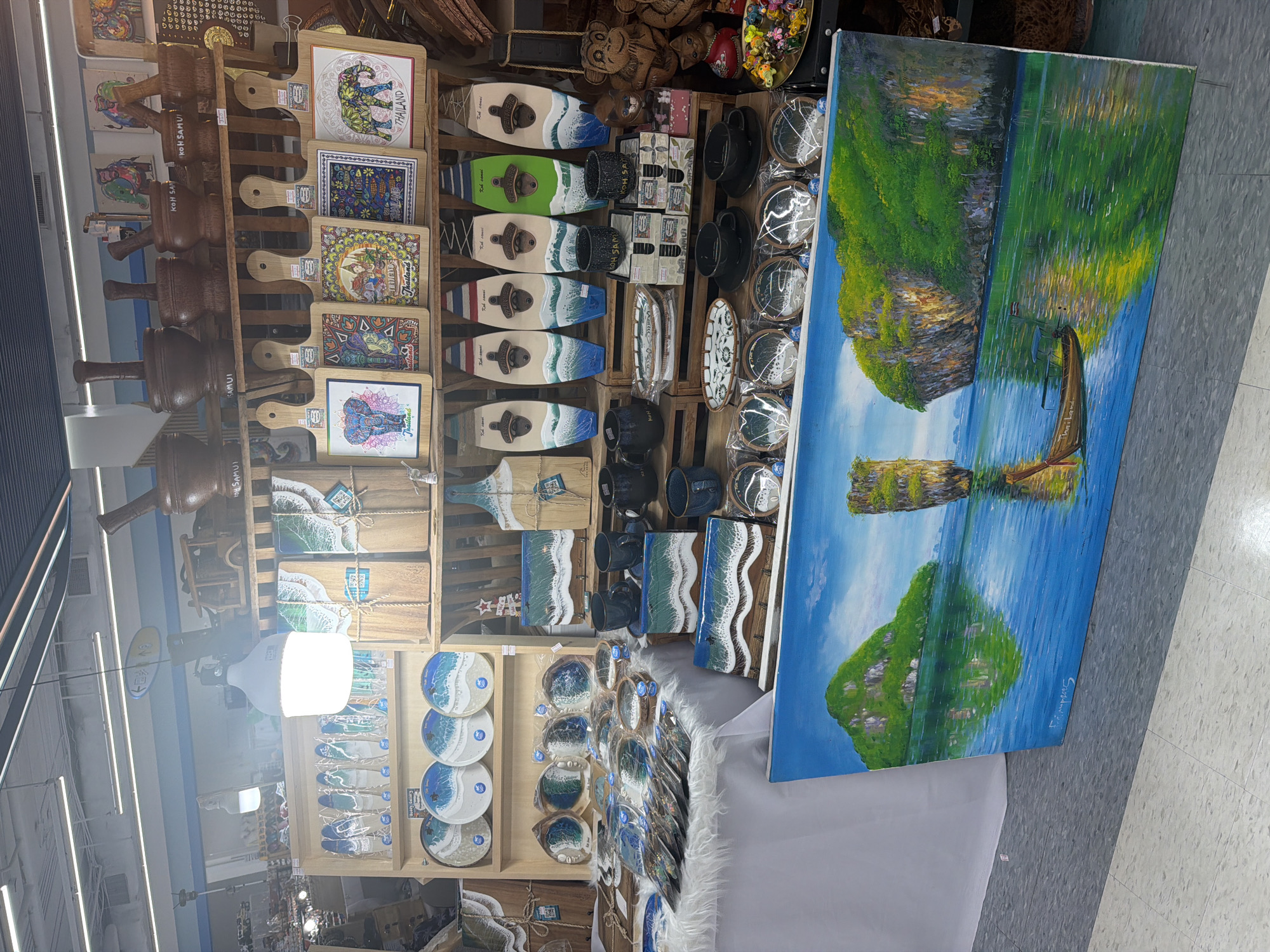 The image shows a well-lit retail display inside a shop or market, likely in บ้านบางรักษ์, Thailand. The display is filled with various artisanal products, many with a coastal theme.
To the left, a vertical arrangement of dark wooden mallets are displayed. Alongside them, decorative cutting boards with elephant and other colorful designs can be seen. Lower down, more cutting boards with resin ocean wave effects are present. Further down are several coasters with ocean wave designs.
To the right of the cutting boards, there are items like coasters, mugs, and surfboard-shaped bottle openers, many with a wave or beach motif. In the foreground, there is a large painting that looks like a seascape with islands, boats, and reflections.
The items are arranged in a grid of wooden shelves. The lighting in the shop is bright, possibly from overhead lamps or recessed lighting. The floor is a light gray tile.