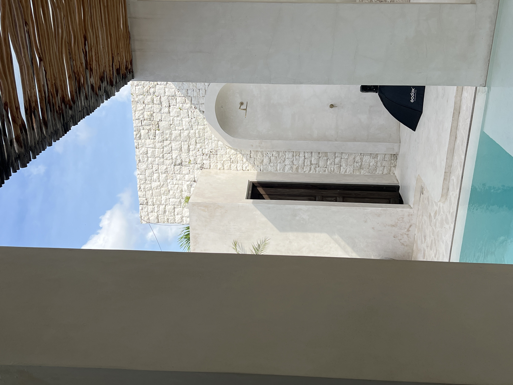 The image captures an architectural view in what appears to be North Kuta, Indonesia, characterized by a blend of modern and traditional design elements. A section of a thatched or woven wood roof is visible in the upper left, contrasting with the smooth, light-colored walls. A patch of blue sky with scattered clouds is framed by the roof and walls. A building with light brickwork, possibly limestone, contains a dark wood-framed window or door. Another building with light concrete-like walls is to the right. The corner of what appears to be a pool with light blue water is present in the upper right of the frame. A black softbox lighting modifier is seen next to the pool, which indicates a photoshoot or a videography set-up. A white wall or ceiling creates a horizontal foreground element, obscuring a portion of the view. Green plants are also seen.