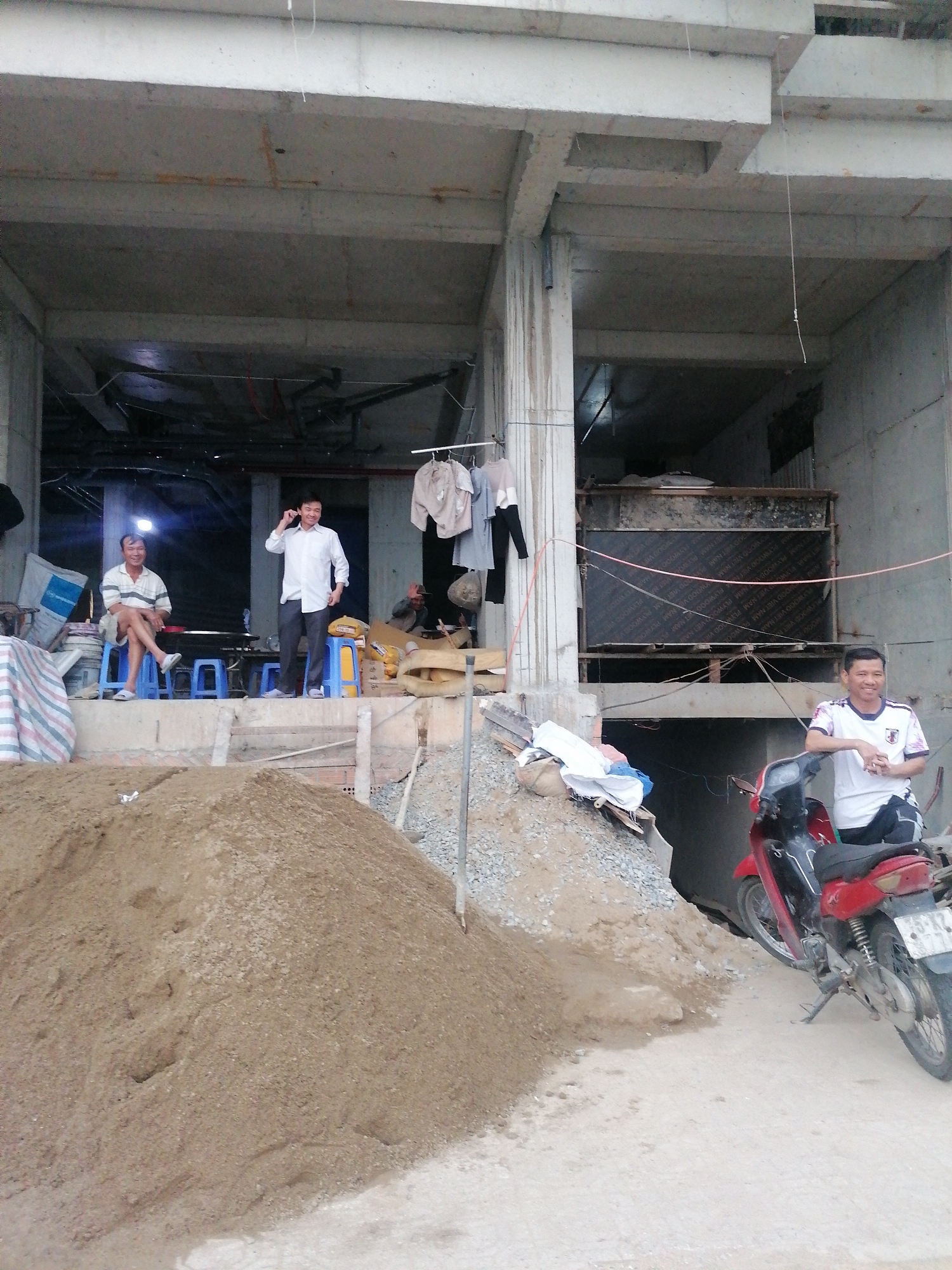 The image shows a construction site in Da Lat, Vietnam. In the foreground, there is a large pile of sand and a smaller pile of gravel. A red motorcycle is parked nearby. In the background, there is an unfinished concrete building. Three men are visible. One man is standing and talking on a phone. Another is sitting on a blue stool. A third man is leaning against the motorcycle. Clothes are hanging on a line near the building's entrance.