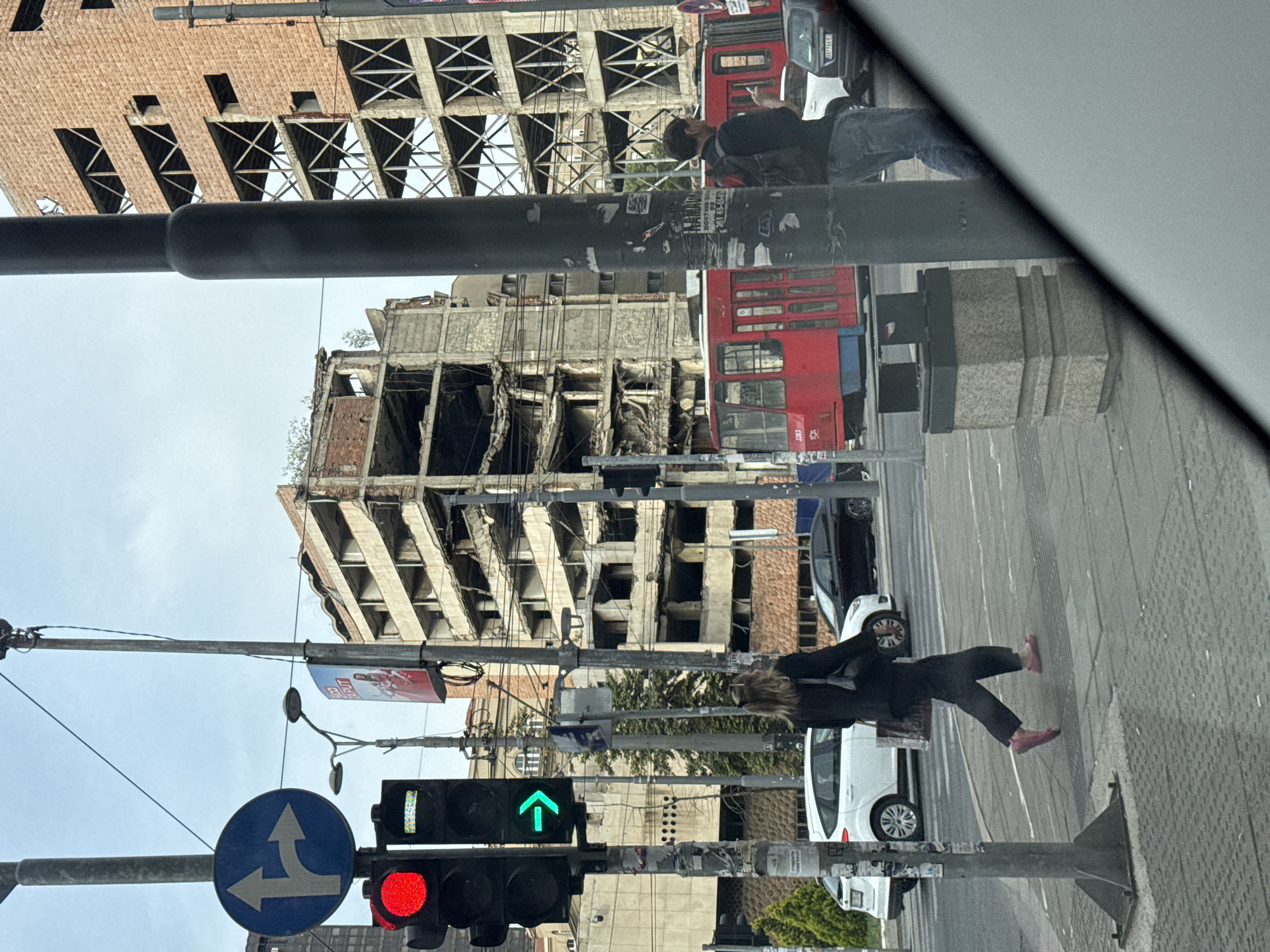 The image depicts a street scene in Belgrade, Serbia, focusing on the bombed-out shell of the former Yugoslav Ministry of Defence buildings. The building’s skeletal structure is visible against the sky. The image is taken from inside a vehicle, possibly a car, looking outwards through the windshield. In the foreground, there is a traffic light showing green for forward movement and red for cross traffic, along with a sign indicating the direction of traffic. A woman is walking on the sidewalk. A red bus and several cars are on the road. A man is walking beside the road. The overall impression is one of urban decay juxtaposed with everyday life.