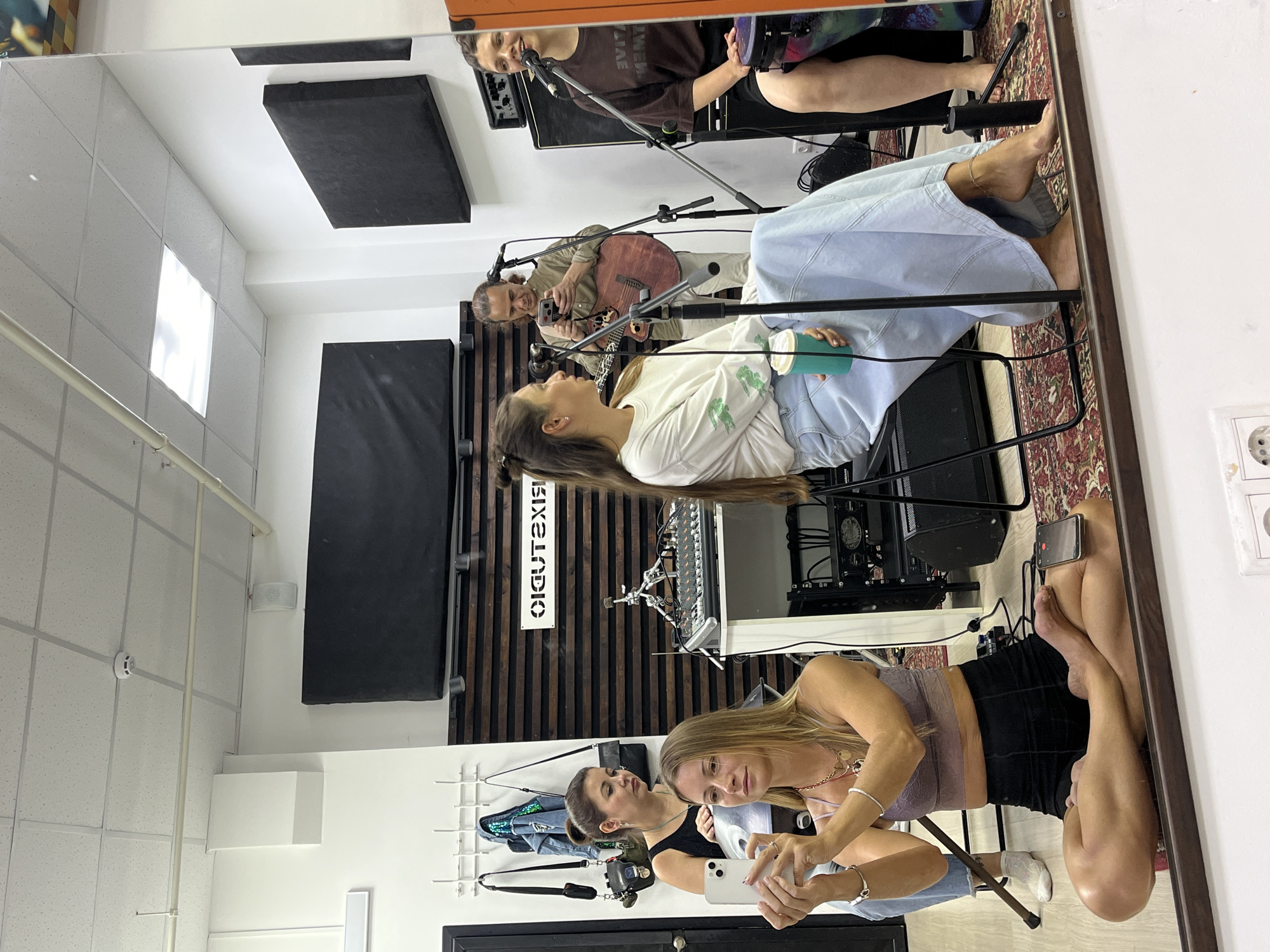 The media file is a vertical, mirrored, full-shot image taken inside a music studio in Moscow, Russia. The scene features four individuals and musical equipment. In the foreground, a woman with blonde hair is seated, holding a smartphone and looking at it. She is wearing a light purple top and a black skirt. To her left, another woman with brown hair is seated, looking to her right. She is wearing a brown t-shirt and black shorts. Behind them, a man with brown hair is seated and playing an acoustic guitar. He is wearing a tan shirt. In the background, a woman with dark hair is standing and singing into a microphone. She is wearing a brown t-shirt. The studio is equipped with soundproofing panels on the walls and ceiling, a mixing board, and various microphones. The lighting suggests it is daytime. The overall mood appears relaxed and creative, with the individuals engaged in making music. Visible text on a sign in the background reads "BIG STUDIO" in a white font on a dark background.