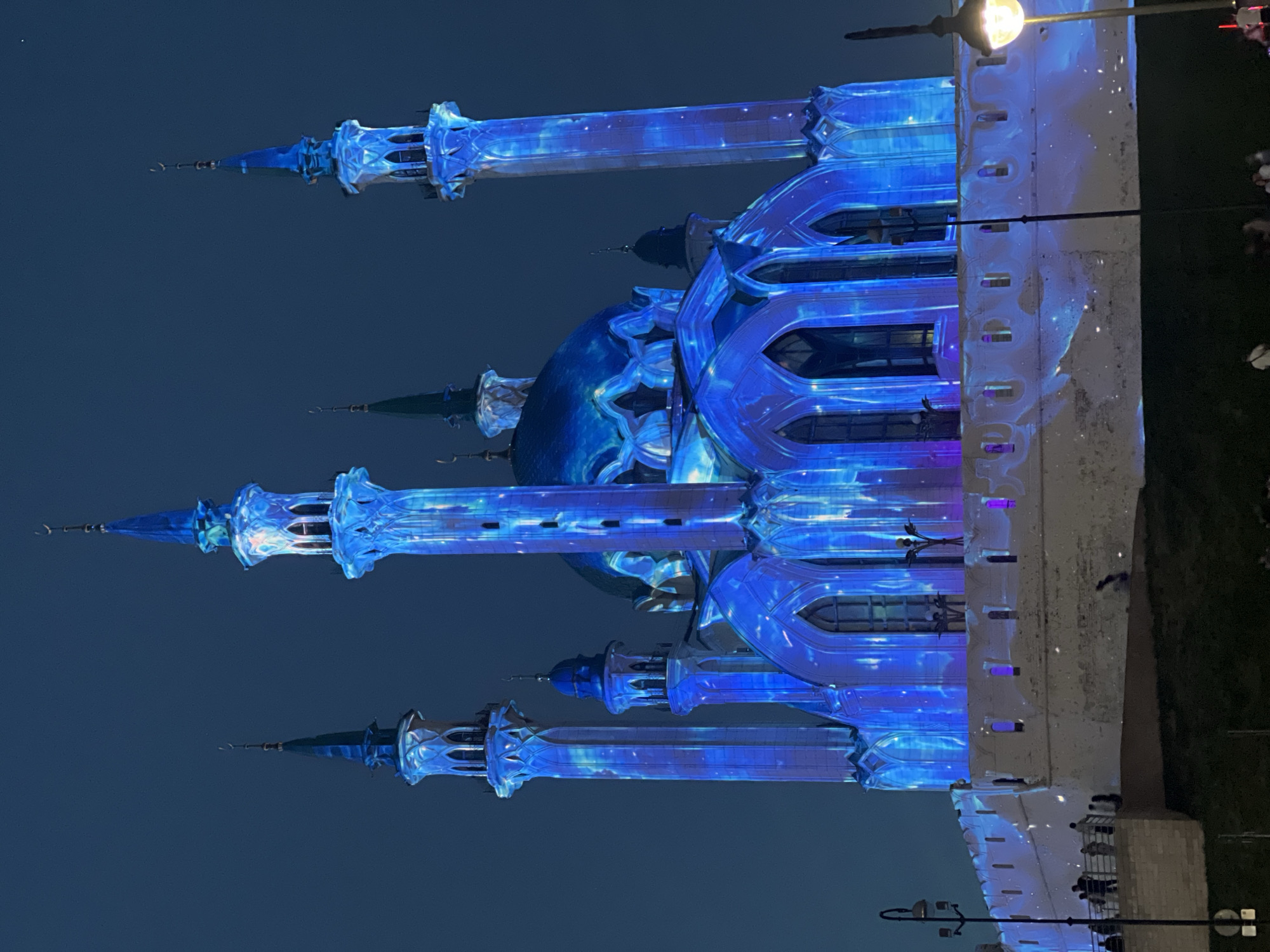 The image shows the Qolsharif Mosque in Kazan, Russia at night. The mosque is illuminated with a blue light display that appears to project nebula-like images onto the building's exterior. The mosque features several minarets and a large central dome. A portion of the surrounding wall is also visible, with a few people standing near it. The sky is dark blue, suggesting it is nighttime. A street lamp is visible in the upper right corner of the photo.