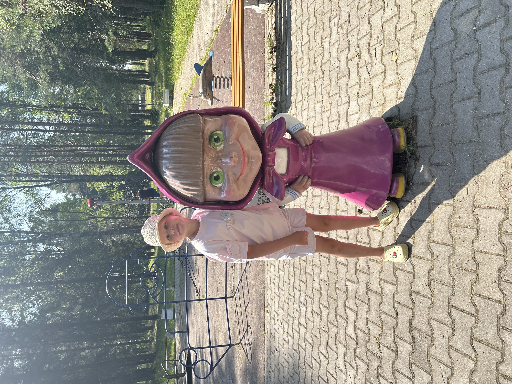 In Krasnovishersk, Russia, a young girl wearing a white bucket hat, a light pink t-shirt, and white shorts stands on a paved pathway in a park. She is posing next to a life-sized statue of a cartoon character, "Masha" from the animated series Masha and the Bear, dressed in a magenta dress. The statue has a large head with wide green eyes and a smiling expression. Behind them, a blue playground structure and trees are visible. In the background, a wooden park bench and a spring-mounted toy are situated near the pathway. The scene is illuminated by sunlight, casting shadows on the ground.