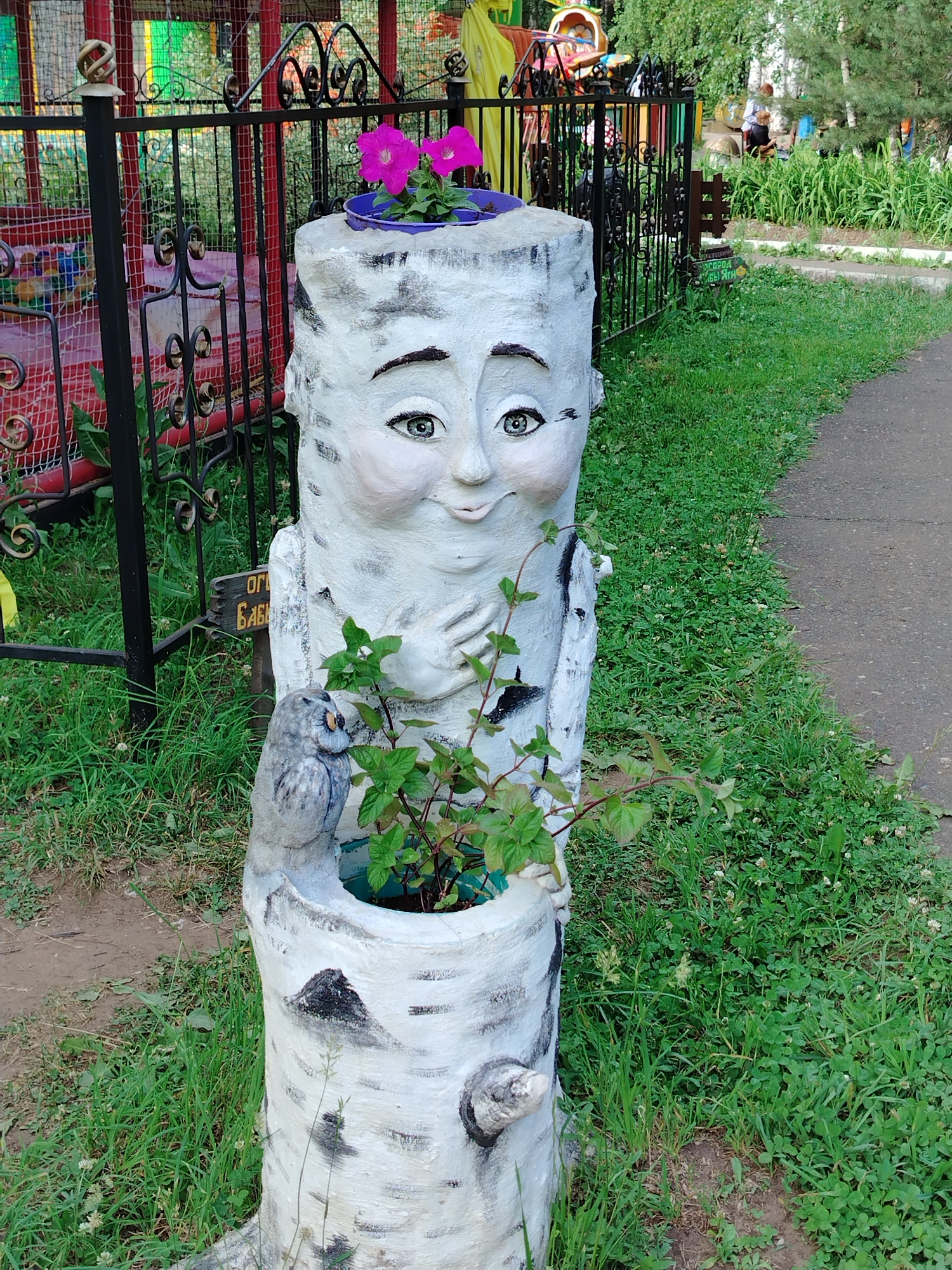 The image shows a concrete sculpture resembling a birch tree trunk with a carved face and an owl figure at its base. The sculpture is located outdoors in a grassy area next to a paved path. In the background, a black metal fence encloses a playground with colorful structures. The sculpture is a planter, with two pink flowers in a pot on its head and a small plant growing from its base. A sign with Russian text is attached to the fence near the sculpture. The environment suggests a public park or recreational area, possibly a children's playground in Chaikovsky, Russia, given the context.