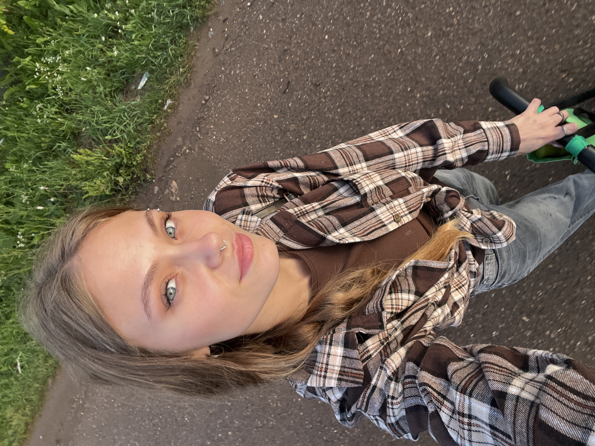 A young woman with blonde hair and blue eyes is captured in a high-angle selfie. She is wearing a brown plaid shirt over a brown t-shirt and blue jeans. Her hair is styled in a braid. She has a piercing on her eyebrow and a nose ring. In her right hand, she is holding the handlebars of a bicycle. The background shows a dirt road with green grass and vegetation on the side. The lighting suggests it is daytime, possibly late afternoon or early evening. There is no identifiable indication of the location being in Fokino, Russia.