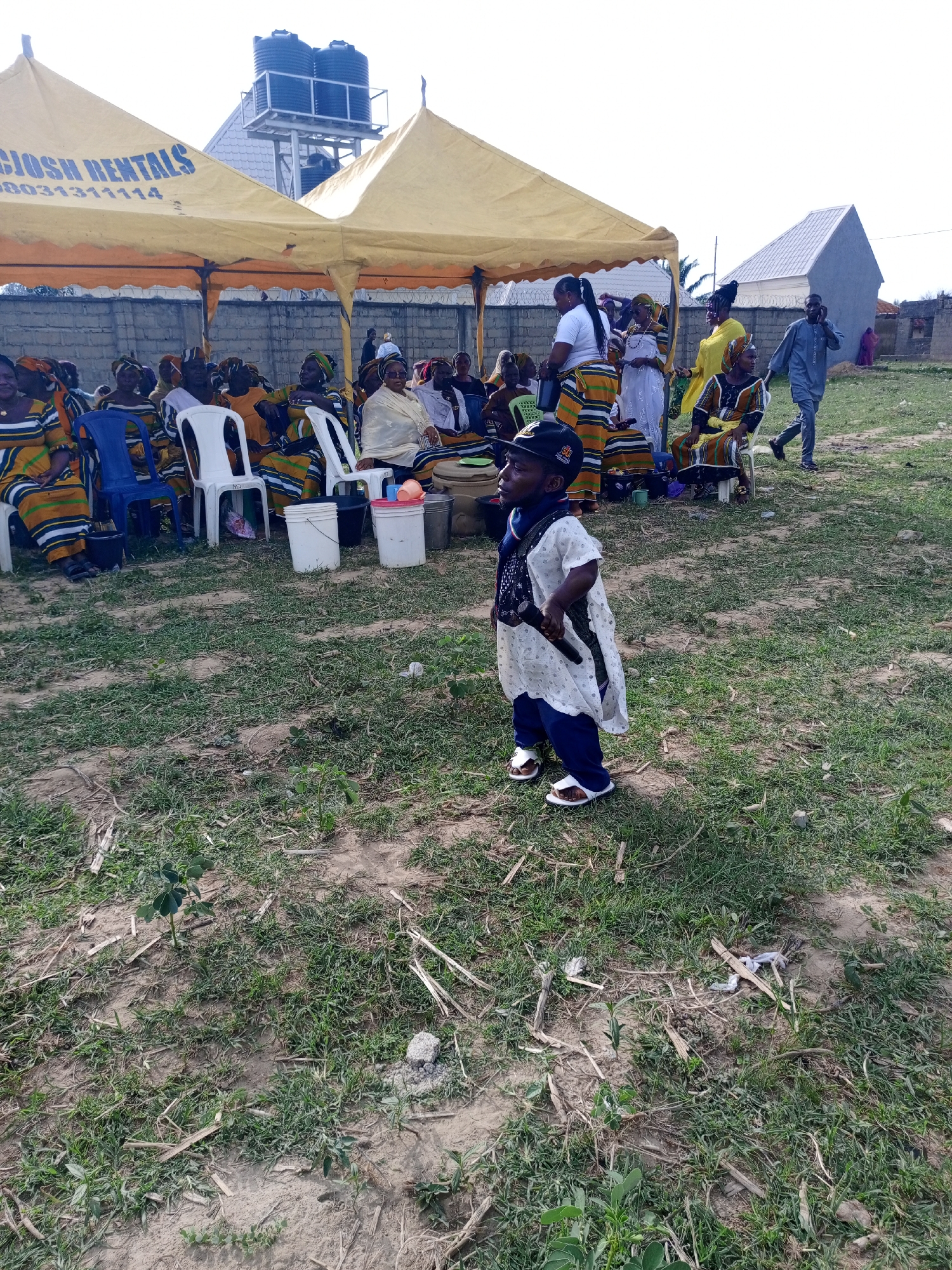 The image depicts an outdoor gathering in Keffi, Nigeria. A small man dressed in traditional attire stands in the foreground on a grassy lawn. Behind him, a group of people are seated under yellow canopies, some wearing colorful, patterned clothing. White plastic chairs are visible among the seating arrangement. In the background, there is a grey building and a water tower with two tanks. A man in a grey outfit walks in the right side background using his phone. There are buckets on the ground with plastic cups stacked next to them. The grass is partially dry and bare patches of earth are visible.