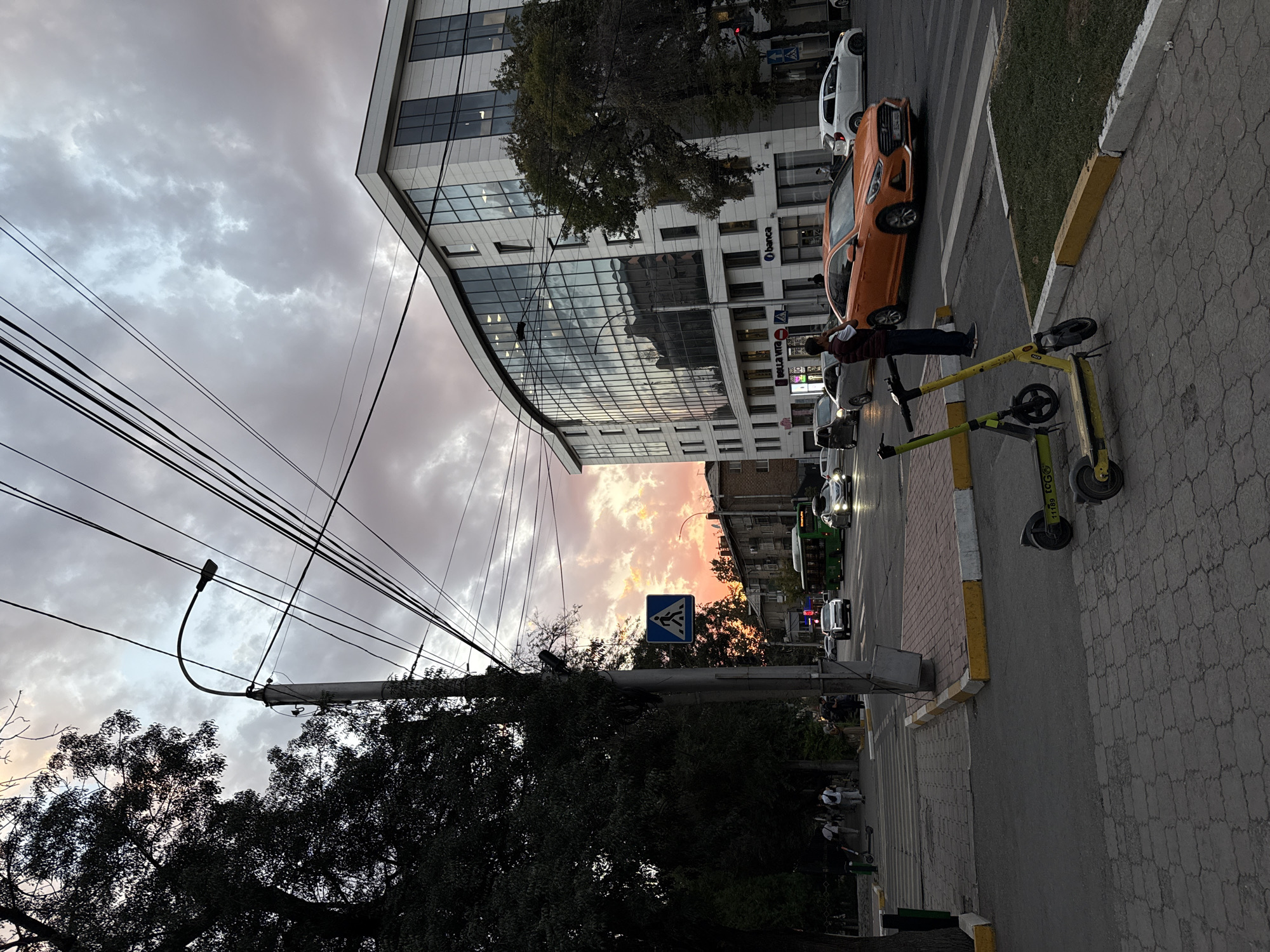 The image depicts a street scene in Bishkek, Kyrgyzstan, during what appears to be dusk or sunset. A modern, multi-story building with a curved glass facade dominates the right side of the frame, housing businesses such as "Banca" and "Bella Vita." Across the street, traffic is visible, including an orange sedan, a white car, a green bus, and other vehicles.
In the foreground, two electric scooters are parked on a paved sidewalk. A person in a maroon vest and dark pants stands near the intersection, looking to their left. The sky above is filled with dramatic clouds tinged with the colors of sunset, casting a warm glow. Power lines crisscross the sky, supported by a lamppost on the left side of the image. Trees line the left side of the street, with some people walking on the sidewalk. A pedestrian crossing is marked by a blue sign with a white pedestrian symbol. The street itself is paved, with a yellow and white striped curb. The overall scene suggests a busy urban environment transitioning into evening.