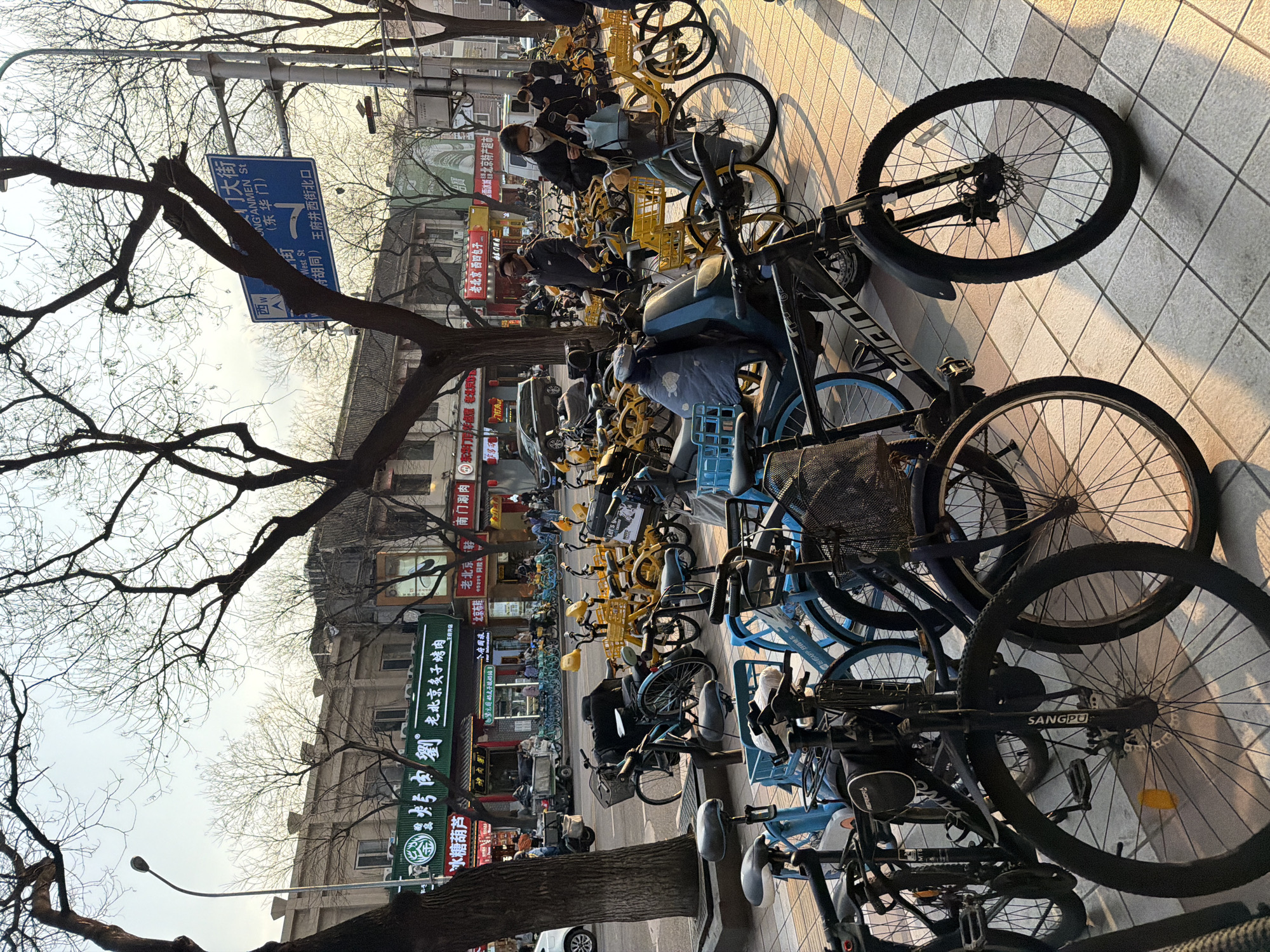 This scene captures a street in the Dongcheng District of Beijing, China. A profusion of bicycles, many of them shared bikes in blue and yellow, line the sidewalk. Interspersed among the bikes are people, some wearing masks. Overhead, bare tree branches reach out against the sky. Buildings line the street, displaying shop signs in Chinese characters. Signage is seen for Wangfujing West Street, and various businesses including a Peking roast meat shop, a hot pot restaurant, and a snow ice city supermarket.