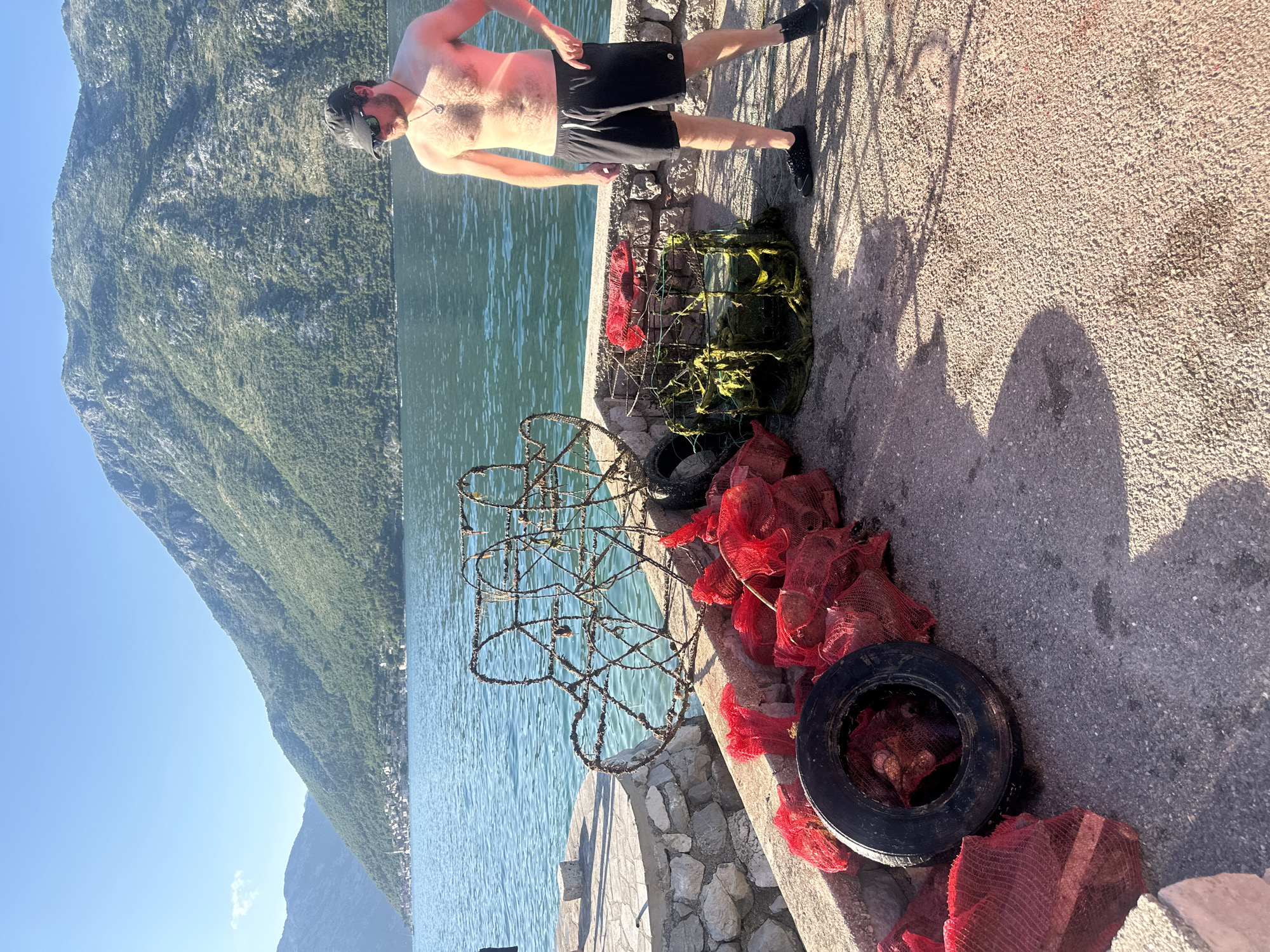 This image captures a sunlit coastal scene in Prčanj, Montenegro. An adult male stands on a concrete pier in the mid-ground, positioned toward the right side of the frame. He is shirtless, wearing black shorts, dark sneakers, a dark baseball cap, and sunglasses, with a pendant visible on a necklace. To his left, on the concrete surface, are various objects: two large, rusty, circular wire-frame structures leaning against each other, a rectangular frame covered in green netting, and a collection of red mesh bags, many containing spherical dark objects. A black rubber tire also rests among the red mesh bags. The pier edge to the left is a low stone wall topped with concrete. Beyond the pier, calm, blue-green water stretches across to the opposite shore, where a town's buildings are clustered at the base of steep, vegetated mountains. The sky is clear blue, with one small cloud visible on the left horizon. Strong sunlight casts distinct shadows across the pier surface.