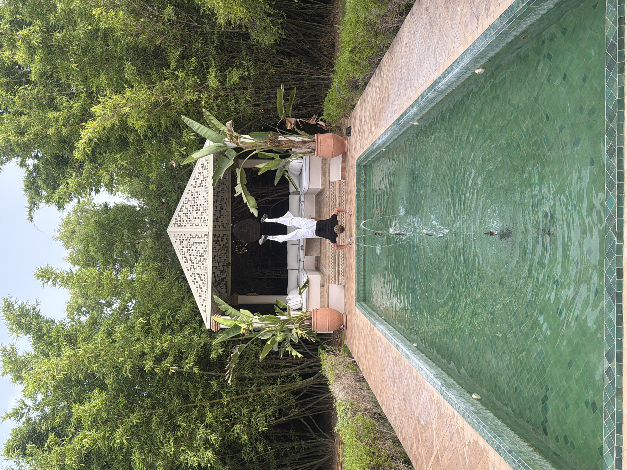 In Al Ouidane, Morocco, a person performs a handstand at the edge of a rectangular swimming pool. The pool has vibrant green tiled walls and a tiled bottom, with a fountain in the center creating ripples on the water's surface. The person is wearing white pants and a dark top, with their legs extended upwards. To the rear of the pool, a pathway of terracotta tiles leads to a white, ornate gazebo. This gazebo features a patterned roof and contains white seating. Flanking the gazebo are two large potted plants with broad leaves, and behind them, a dense wall of tall bamboo stalks forms a natural backdrop. The area is bathed in natural daylight.