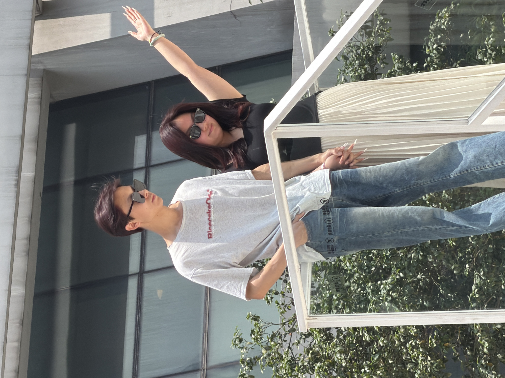 A young man and a young woman stand on a white railing of an outdoor staircase, holding hands. The man, on the left, wears a light grey t-shirt with red lettering that reads "RicecakeObs," sunglasses, and blue denim jeans. The woman, on the right, wears a black top, a cream-colored pleated skirt, and sunglasses. Her left arm is raised with her hand extended upward. They are positioned against a backdrop of a modern building with large, dark windows and a lush green tree. The location is Yuzhong District, China.