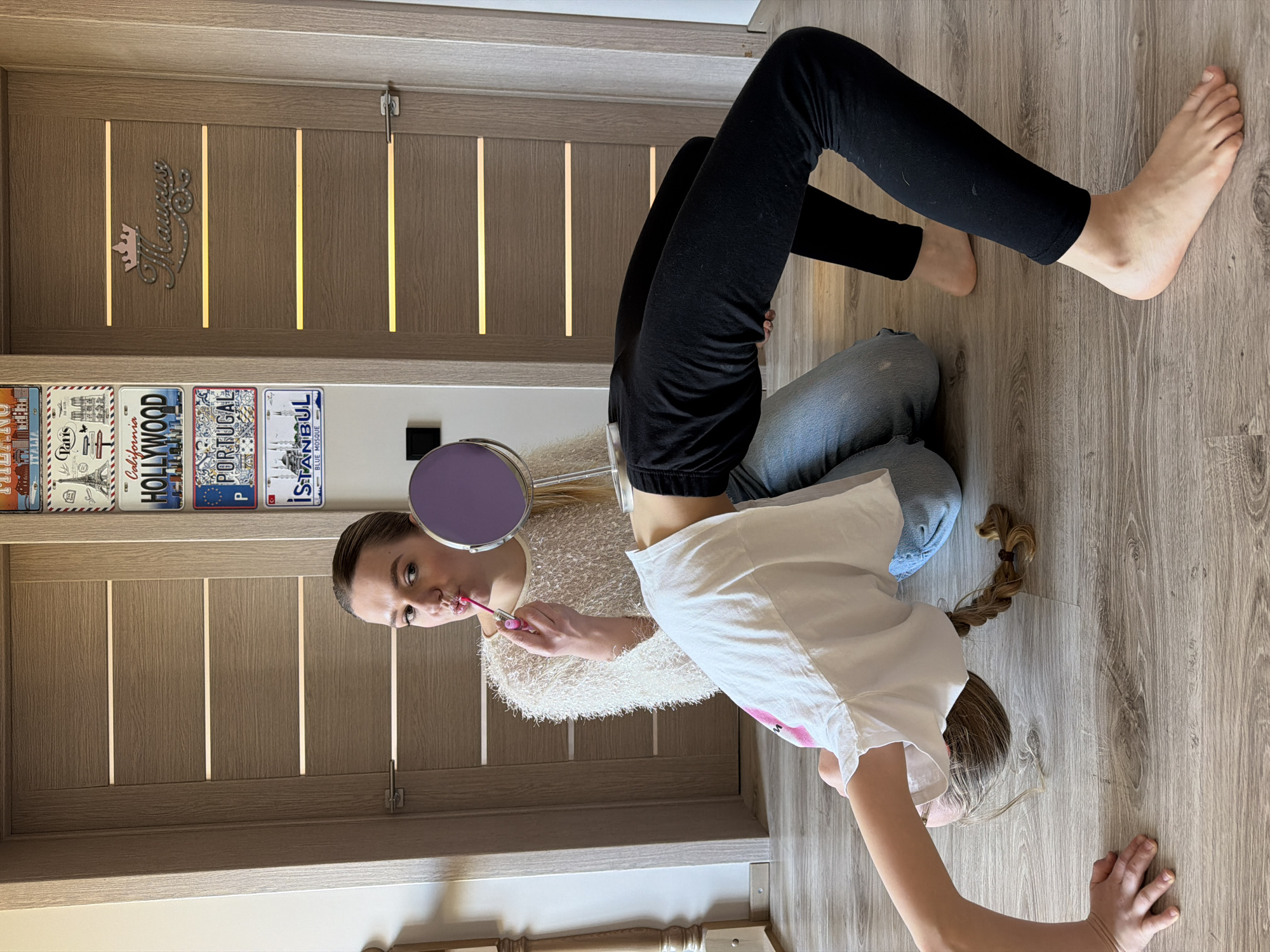 An indoor, brightly lit scene in Dreiliņi, Latvia, shows two females in a domestic setting, likely a home. The image is oriented vertically with the floor to the left and a door to the right.
One female, appearing older, is positioned on her knees and bent backwards, looking into a small, round, purple-framed mirror held in her left hand. She wears a fluffy, light-colored (possibly white or cream) top and black leggings. With her right hand, she applies pink lip gloss from an applicator to her lips. She has a focused expression.
Below her, a younger female lies face down on a light wooden floor, wearing a white short-sleeved t-shirt and blue jeans. Her light brown hair is in a braid that rests on the floor. Her face is partially obscured as she looks down, with her right arm extended forward on the floor.
The background features a light wooden door with embedded vertical light strips and a silver, crown-like decoration with the name "Margarita" above it. To the left of the door, a wall is adorned with several decorative license plates from various locations, including "Paris", "California HOLLYWOOD", "PORTUGAL", and "ISTANBUL BLUE MOSQUE". The flooring throughout the visible area is a light wood laminate. The overall impression is a casual, playful moment.