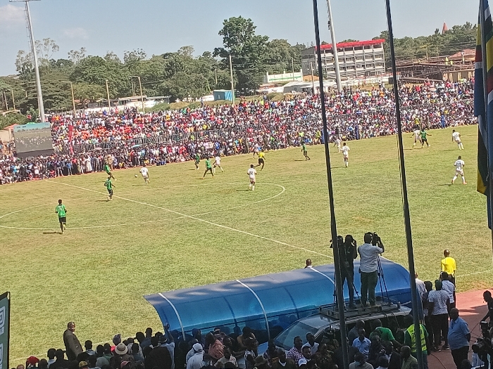 The image depicts a lively daytime football (soccer) match taking place in a crowded stadium, likely in Siaya, Kenya. Hundreds, if not thousands, of spectators fill the stadium stands, creating a vibrant atmosphere. The stands are packed with people, mostly dressed in casual attire, indicating a significant local event.
On the lush green pitch, players from two teams are actively engaged in the game. One team wears white jerseys with numbers (e.g., player #11 and #13 are visible), while the opposing team is dressed in green jerseys. The football is visible near the center of the field, with several players converging around it, suggesting a moment of active play.
In the foreground, on the right side, two cameramen are positioned on a raised platform or vehicle, diligently filming the match, indicating a professional broadcast or recording of the event. Below them, a blue-roofed dugout or shelter is visible, with more spectators and officials gathered around.
The match is taking place under a clear, bright blue sky with abundant sunshine, casting distinct shadows on the field and creating strong illumination. In the background, beyond the stadium, there are several green trees and multi-story buildings, suggesting an urban or semi-urban setting. Partially visible flagpoles on the right display flags with stripes that appear to be yellow, black, and red, which could be indicative of the national flag of Kenya or a neighboring country. The overall scene captures the excitement and energy of a community sporting event. A barely discernible green logo on an advertising board on the far left edge of the pitch is too distant to read.