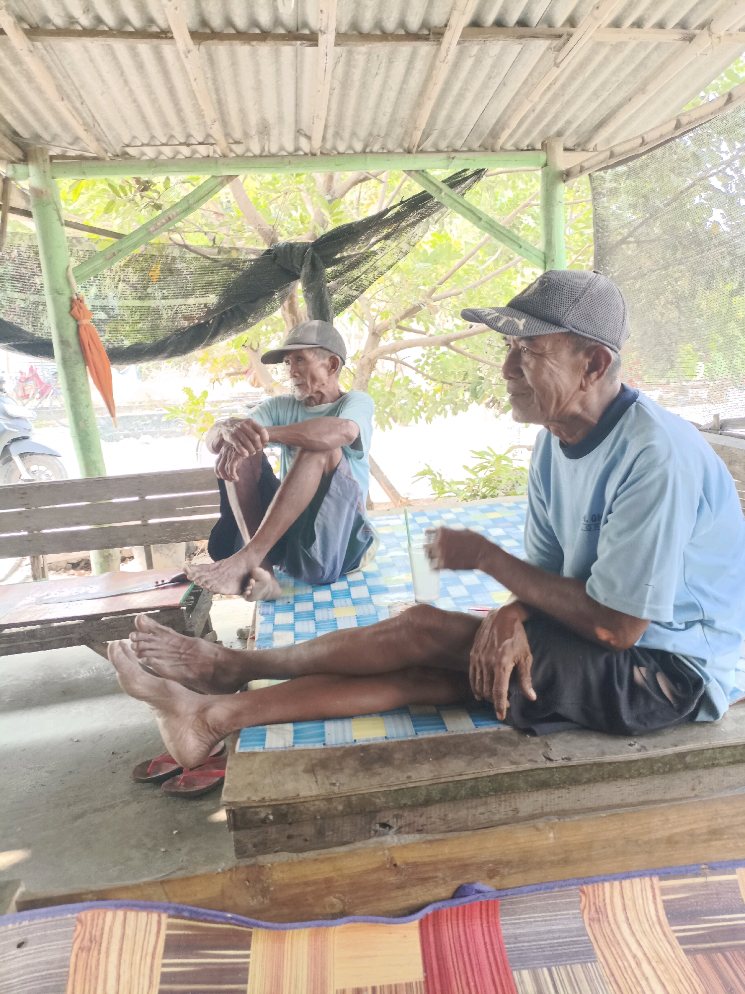 Two elderly men are seated outdoors under a corrugated tin roof supported by bamboo poles. The man on the right, facing left, wears a light blue t-shirt and dark shorts, and a grey baseball cap. He holds a glass with a straw in his right hand. The man on the left, facing right, also wears a light blue t-shirt and dark shorts, and a grey baseball cap. He is seated with his legs crossed and his arms resting on his knees. Both men appear to be in a casual setting, possibly a rest area or a simple dwelling. In the background, there are trees and foliage, suggesting a rural or semi-rural environment. A motorcycle is visible to the left, parked near the entrance of the shaded area. The ground is concrete, and a colorful mat is laid out on the floor in the foreground. The overall impression is one of a peaceful, everyday scene.