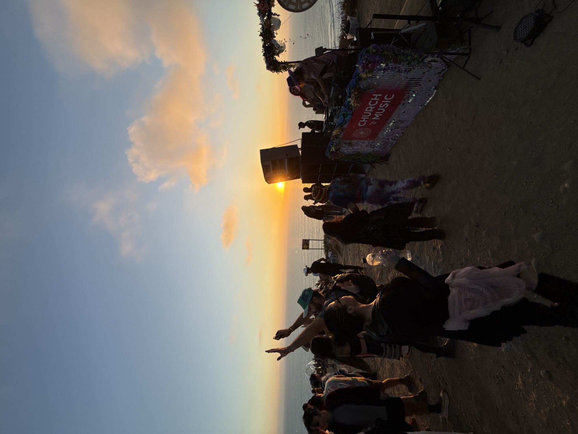 The image shows a sunset gathering on a beach in San Diego, California. A crowd of people are silhouetted against the setting sun over the ocean. A DJ booth decorated with "Church of Music" signage is set up at the edge of the beach. Speakers are positioned to amplify the music, and some decorations adorn the booth. The sky displays a gradient of colors from blue to orange, with a few scattered clouds. The sand covers the foreground. People are facing the ocean.