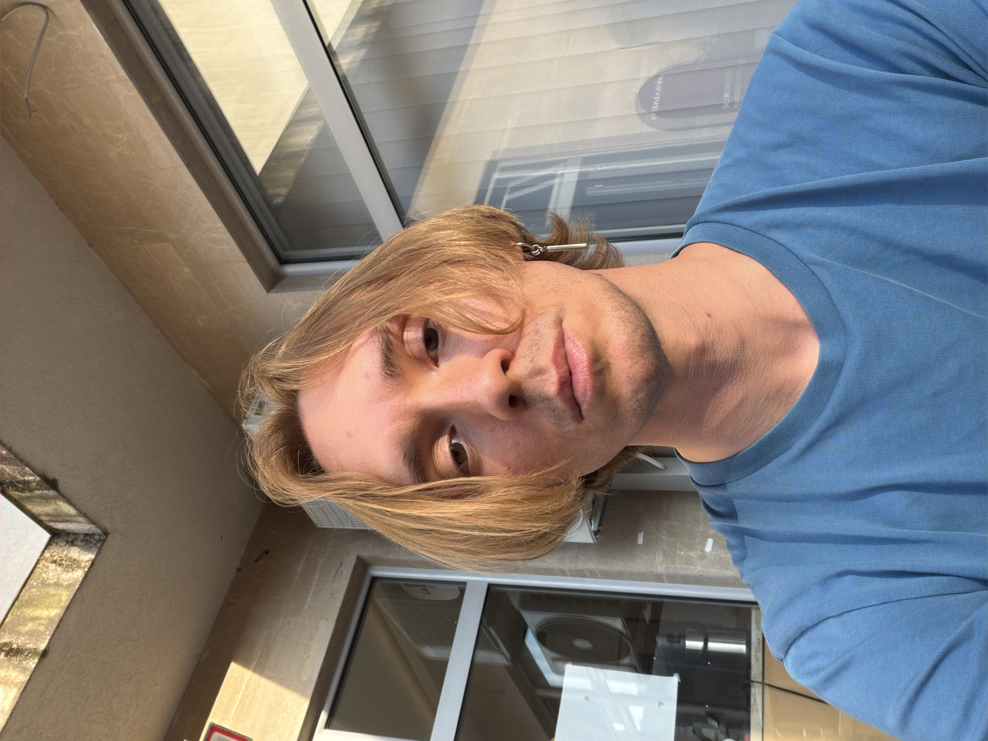 A rotated close-up portrait features a young man with medium-length, light brown hair and light facial stubble, wearing a blue t-shirt. He looks directly at the camera with a neutral expression. A silver-toned, bar-shaped earring is visible in his left ear.
The man is positioned in what appears to be an outdoor or semi-outdoor urban setting, possibly a balcony or patio, adjacent to a building. Behind him, a large window reflects strong, angular shadows, indicating bright daylight. The reflection also shows an air conditioning unit or fan, and faint, inverted, illegible text from an unknown source. To the left, a light-colored wall or tiling with a visible grey cable is present. A lower window frame shows signs of weathering or dirt. The scene conveys a sunny day in an urban environment, consistent with locations such as Budva, Montenegro.