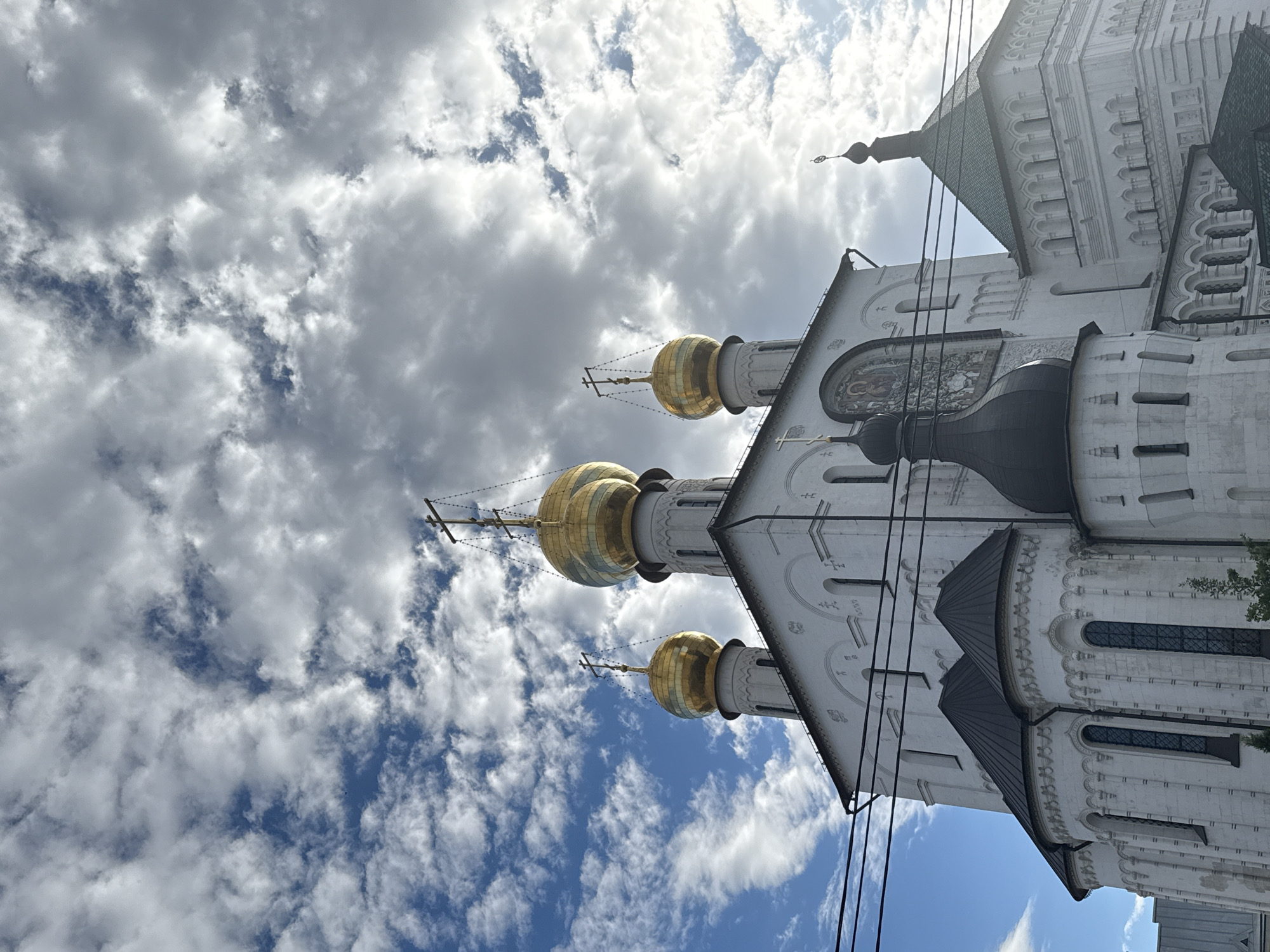 This is a low-angle, full shot of the Trinity Cathedral in the Alexander Nevsky Lavra, Saint Petersburg, Russia. The sky is a mix of blue and dramatic, white and gray clouds, with sunlight breaking through in places. The white, stone cathedral is the dominant subject, featuring multiple golden domes topped with crosses. The domes are arranged in a cluster, with four prominent ones visible in the foreground and a smaller one to the right. The architecture is detailed, with arched windows and decorative elements on the facade. A dark, conical dome with a cross is positioned in front of the larger domes. The lower portion of the building shows more detailed stonework and arched openings. In the foreground, several black power lines are strung across the frame, partially obscuring the view of the cathedral. The scene is devoid of people and animals. The overall impression is one of grandeur and religious significance, set against a dynamic sky.