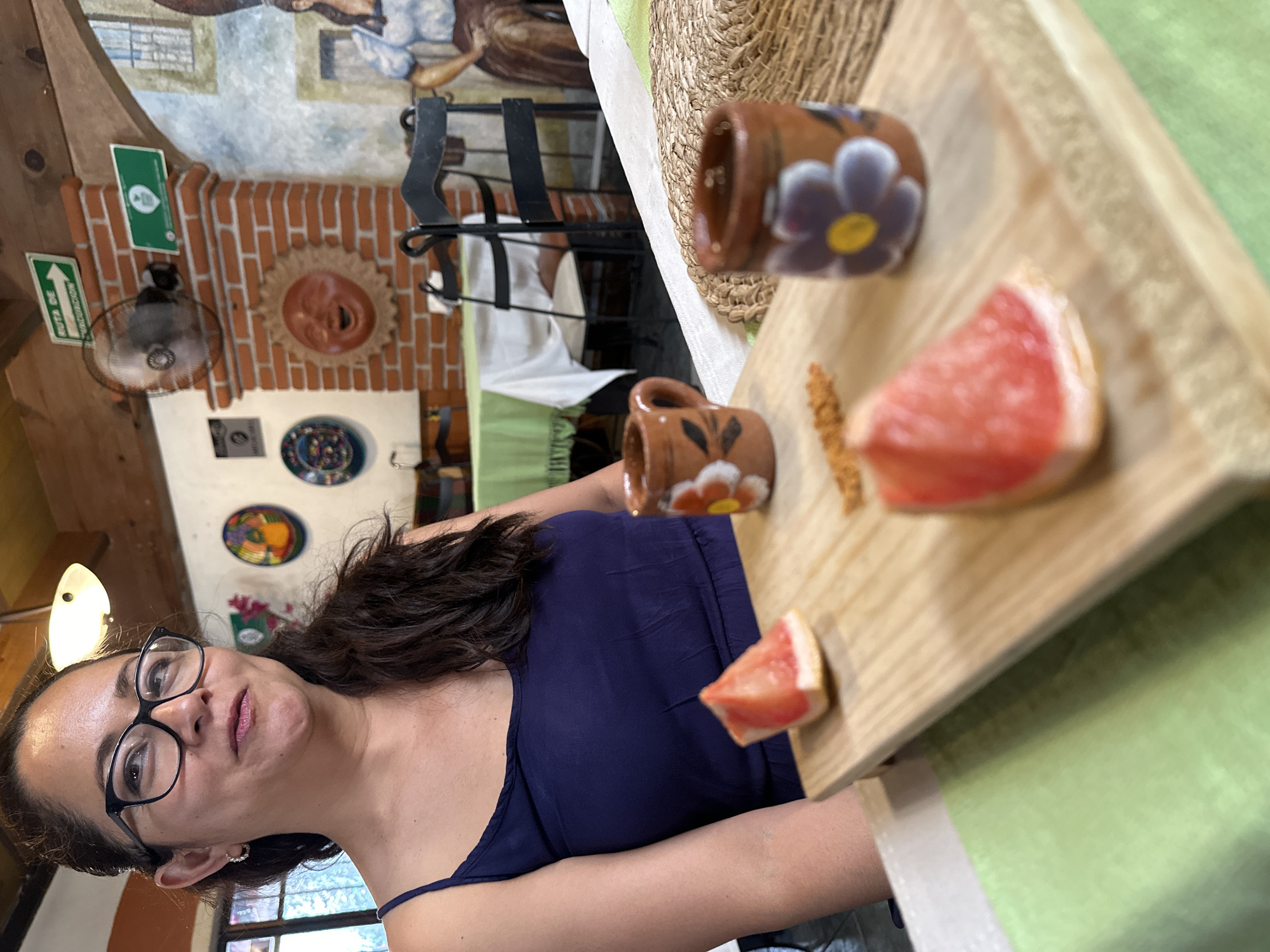 The image depicts a woman with dark, wavy hair and glasses, wearing a dark blue tank top, seated at a table in what appears to be a restaurant. She is holding a wooden board on which there are two small, brown, flower-decorated cups, pieces of pink grapefruit, and some brown granules. The restaurant's interior has a rustic feel, with a brick wall featuring a sun-shaped decoration, plates on the wall, and a painted mural. There's a green exit sign and a fan visible on the wall. Based on context clues, the location is in the city of Puebla, Mexico.