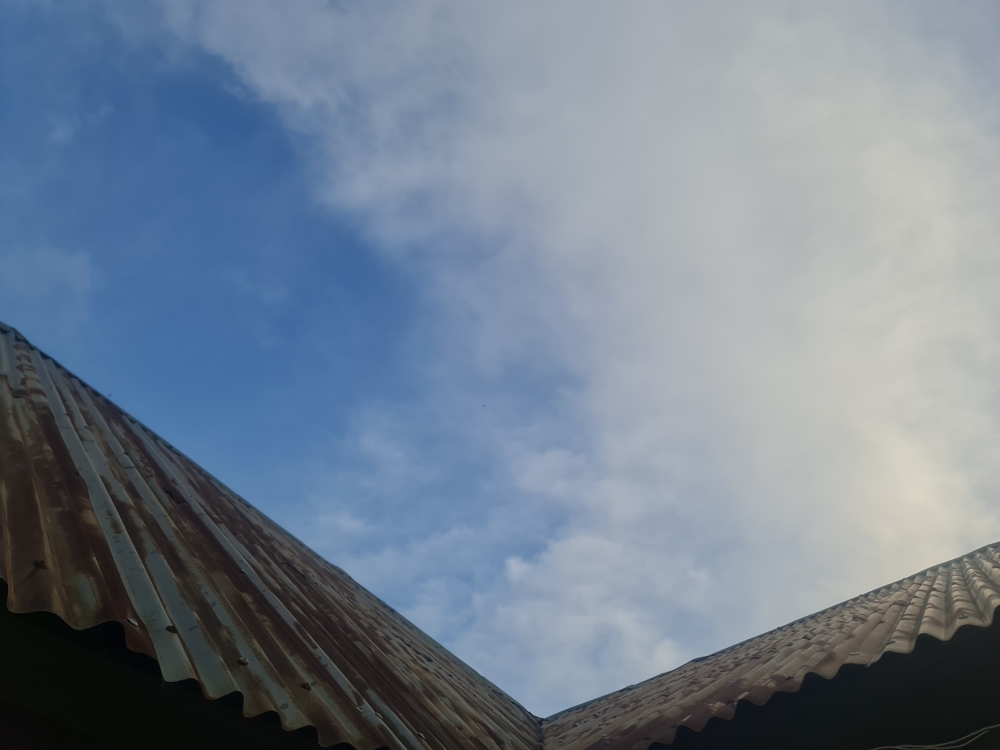 An upward-angle shot of two sections of a corrugated metal roof meeting at a peak, set against a sky with scattered white clouds and patches of blue. The roof sections are weathered and appear to be made of rusted metal. The sky is brightest towards the right, suggesting the sun may be in that direction. The location is the city of Kurunduma, Nigeria.