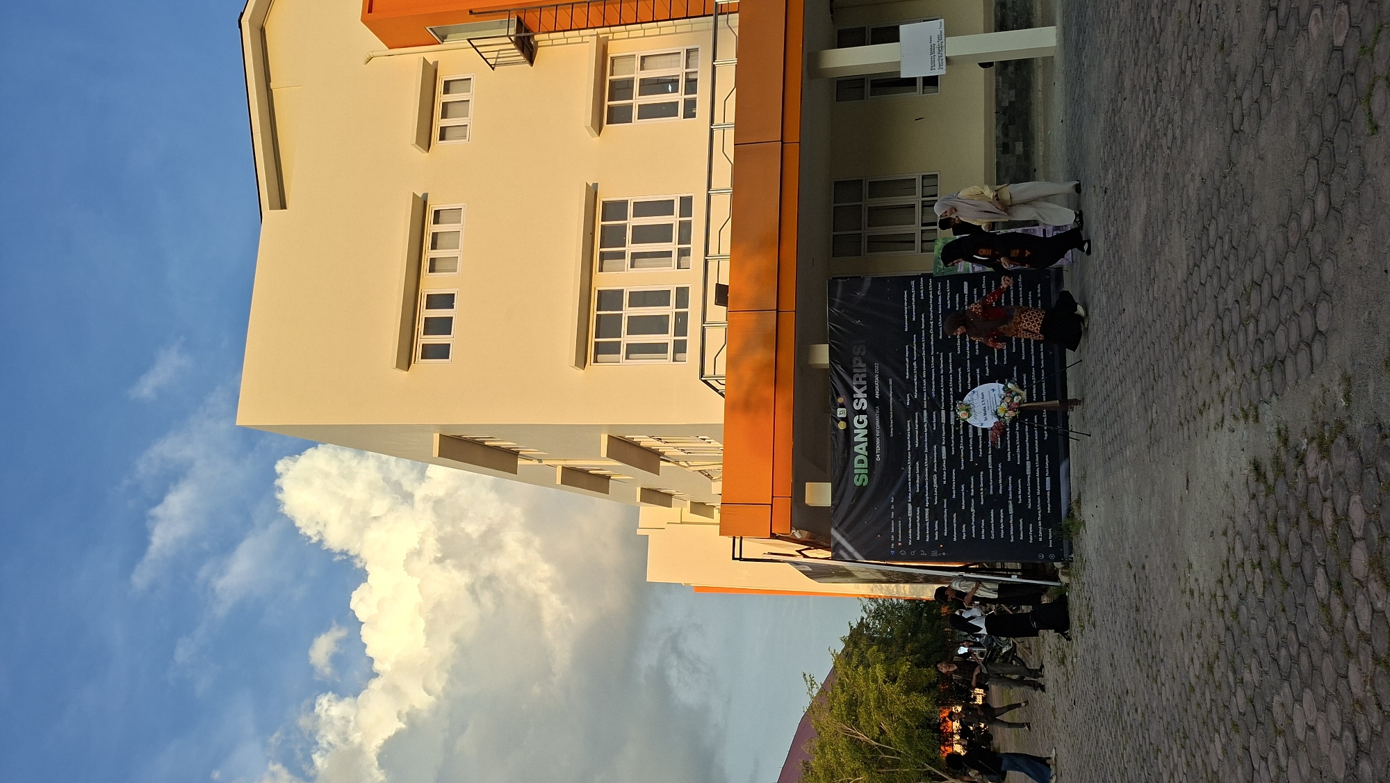 An outdoor daytime scene features a multi-story building with cream-colored walls and orange accents. The building has numerous windows and a fire escape. In the foreground, a large black banner with white text is displayed, listing names under the heading "SIDANG SKRIPSI D4 TEKNIK INFORMATIKA ANGKATAN 2022." Several people are gathered around the banner and in front of the building. Some individuals are standing and conversing, while others are walking. A wreath rests on a stand in front of the banner. The ground is paved with interlocking concrete blocks. The sky is partly cloudy with bright blue sky visible between fluffy white clouds. The location is the Faculty of Engineering, Universitas Malikussaleh, in the city of Lhokseumawe, Aceh Province, Indonesia.