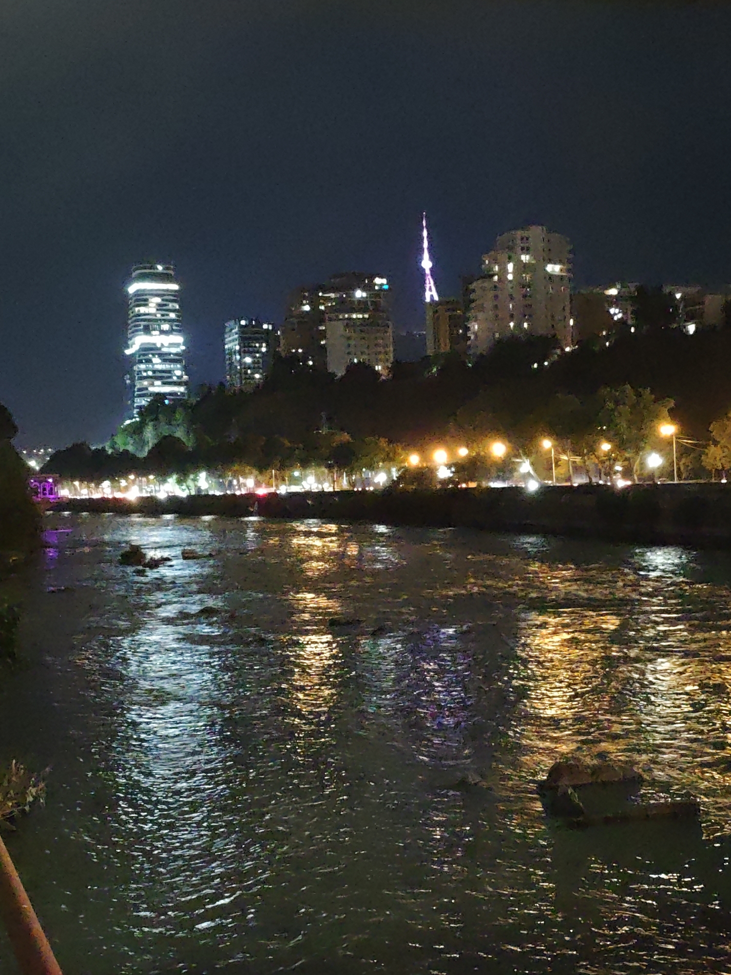 The image depicts a night scene of the Mtkvari River in Tbilisi, Georgia, with reflections of city lights on the water's surface. On the far bank, a cityscape features illuminated buildings, including a tall, modern skyscraper on the left and a broadcast tower with a glowing spire further to the right. Trees and streetlights line the riverbank, casting a warm glow that is mirrored in the water. The river itself appears somewhat turbulent, with ripples disturbing the reflections. The overall impression is one of a vibrant urban environment at night, viewed from across the river.