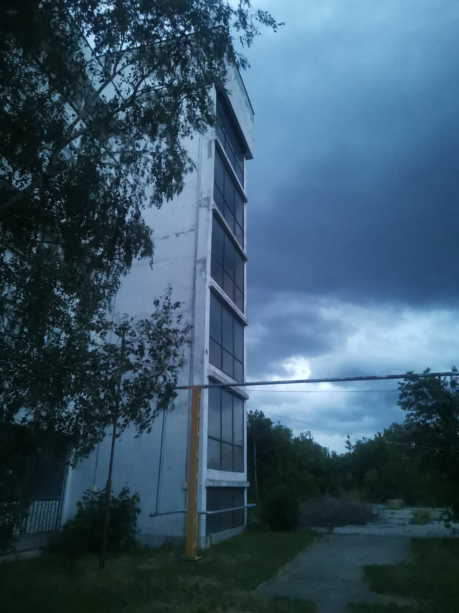 This vertical shot captures a somber scene, likely in Zhivkovo, Bulgaria, featuring a multi-story building under a dramatic, overcast sky. The building, made of a light-colored material, possibly concrete, stands on the left side of the frame, showcasing several dark, rectangular window panes or panels arranged vertically.
Dense, dark green foliage from a tree largely obscures the left facade of the building, with its branches extending across the upper left. A distinct bright yellow vertical structure, possibly a pipe or support, is visible at the base of the building. A horizontal metal pipe or conduit spans the mid-ground, extending from the building towards the right.
The sky, dominating the right and upper portions of the image, is heavily clouded, exhibiting a deep, dark blue at the top that lightens to greyish-blue tones with visible cloud masses closer to the horizon. This suggests stormy weather or the approach of a storm, contributing to a melancholic and muted atmosphere. The lighting indicates late afternoon, dusk, or early evening.
Below the building, there is an expanse of green grass and a grey, somewhat uneven path or ground surface. In the background, a dense line of trees hints at a natural or untamed environment beyond the immediate vicinity of the building. No people are visible, and no specific activities are taking place. The overall impression is one of quietude and a slightly desolate or neglected setting. There is no visible text in the image.