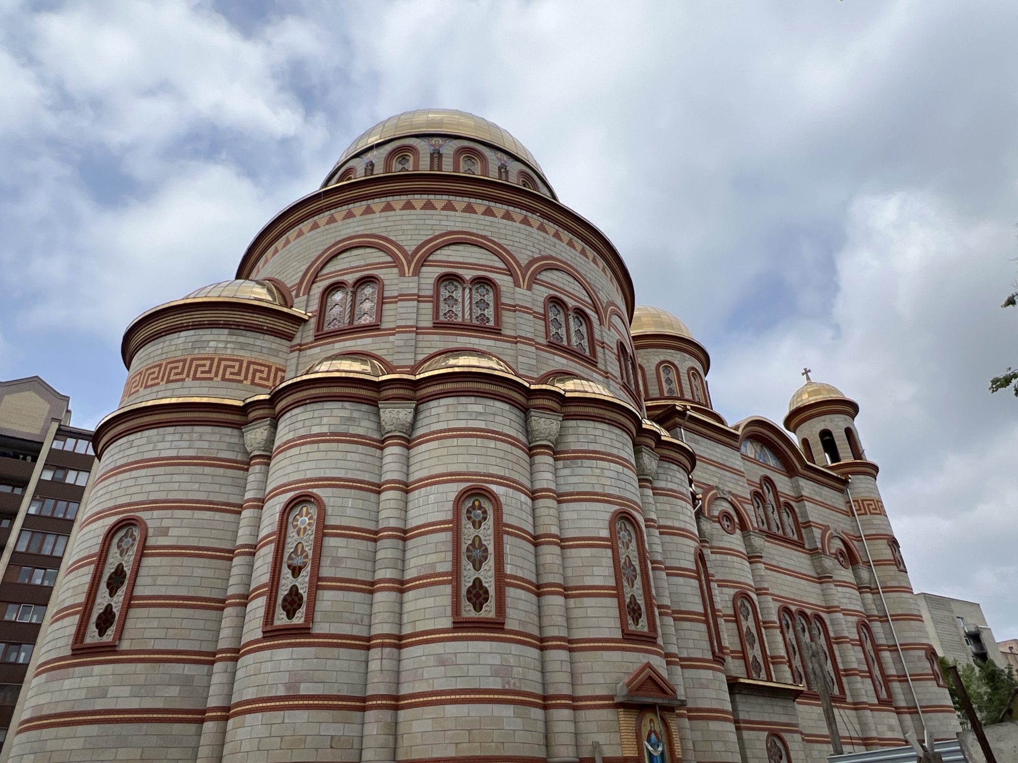 The image showcases a magnificent, ornate church or cathedral, likely located in Ternopil, Ukraine, given the context. The architecture is characterized by light-colored, possibly grey, stone or brick walls laid in horizontal courses, accented with prominent reddish-brown stripes and decorative bands. A distinctive golden Greek key pattern is visible along several architectural elements, adding a classical flourish.
The building is adorned with multiple gleaming golden domes of varying sizes, with the largest central dome rising majestically. Several of these domes are topped with golden Orthodox crosses. The facade features numerous arched windows, many of which contain intricate decorative grilles or possibly stained glass, enhancing the building's detailed aesthetic. Pillars and decorative friezes further embellish the structure, contributing to its grand appearance. On the right side of the building, a smaller bell tower with its own golden dome and cross is visible. Near the base of the church, a small religious icon or painting, possibly depicting the Madonna and Child, is set into a niche.
The photograph was taken on an overcast day, with a sky full of soft, diffused clouds that highlight the golden domes and the intricate details of the stone and brickwork. In the background to the left, a modern, multi-story building, contrasting with the church's traditional design, can be seen. Some greenery and what appears to be a fence or barrier are visible towards the bottom right. The upward angle of the shot emphasizes the impressive scale and grandeur of the edifice. There are no people visible, and no discernible text other than the decorative patterns.