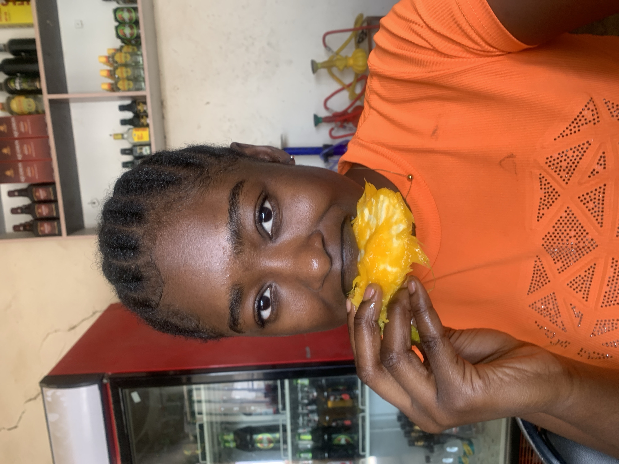 A woman with dark skin and braided hair, wearing an orange top with sparkly designs, is holding and eating a yellow fruit. The background shows shelves stocked with bottled goods, likely beverages or liquor, and a red refrigerator with glass doors displaying various items inside. The location is likely in Kabong, Nigeria, judging by the question's prompt.