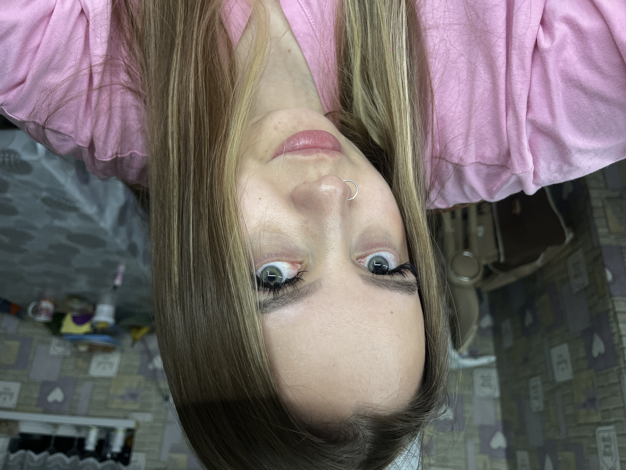 A portrait of a woman with blonde hair, wearing a pink shirt and a nose ring, oriented upside down. The background appears to be a room with a tiled floor and various objects scattered on a shelf. No location information pertaining to Port-Katon, Russia, is present.