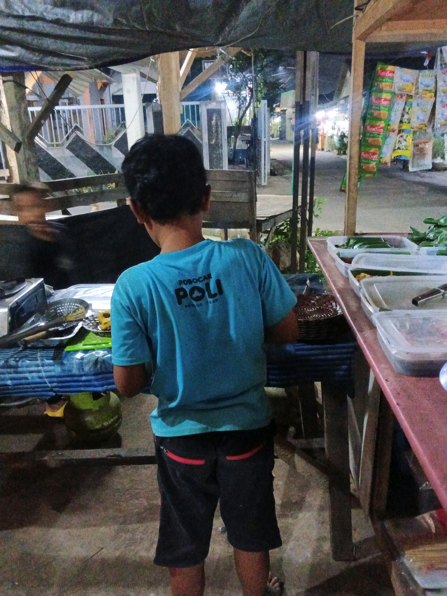 The image depicts an outdoor food stall in Kadu, Indonesia, possibly at night given the lighting conditions. A young person wearing a blue t-shirt with "ROBOCAR POLI RESCUE TEAM" printed on the back and black shorts with red trim is standing with their back to the camera. They appear to be interacting with or observing the food being prepared or displayed.
In the foreground, a blue and white striped tarp covers a table with various food items. A silver cooking utensil with a perforated head is visible, along with a green cutting board and a stack of round, dark-colored food items on a plate. To the left of this table, a round, green object, possibly a coconut or a large gourd, is partially visible.
To the right of the image, a long, reddish-brown table displays several clear plastic containers filled with food, mostly green leafy vegetables, and what appear to be portions of cooked food. Some of these containers are covered. Behind these tables, there are wooden structures and hanging items that appear to be promotional materials or merchandise, some of which are brightly colored and feature text.
The background reveals a pathway or street with more stalls and structures, illuminated by artificial lights. Trees and foliage are also visible, suggesting a relatively informal outdoor market or street food setting. The overall impression is that of a bustling, albeit dimly lit, local food vendor area.