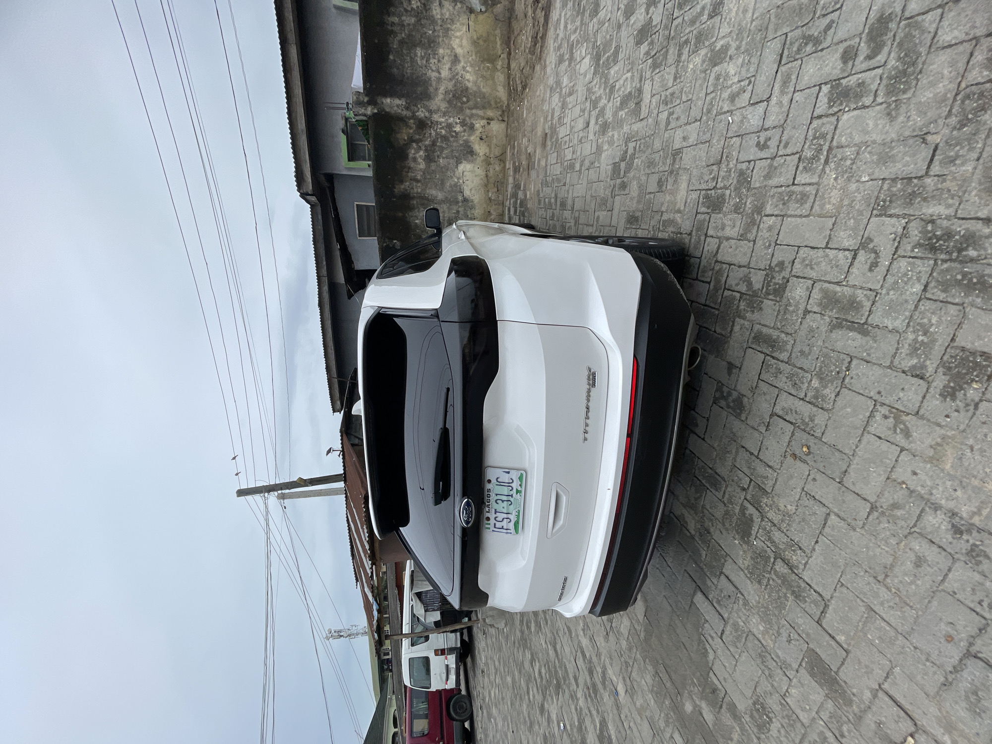 The image shows the rear of a white Ford Edge parked on a gray brick pavement. The vehicle has a Nigerian license plate that reads "FST-31JC," indicating Lagos State. Background details include a building with a corrugated metal roof, a gray wall, and utility poles with electrical wires. Another vehicle or vehicles are partially visible behind the Ford Edge, including what seems to be a white van. The scene appears to be in a residential or commercial area. There is no clear evidence pinpointing this exact location in Ado, Nigeria.