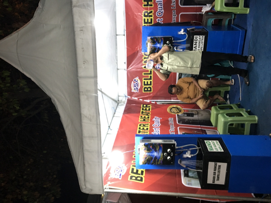 The image shows a booth in Kecamatan Batu, Indonesia, at coordinates (-7.866, 112.514). The booth features two water heaters displayed on blue stands with black trays and black boxes at the base. The booth is located under a white tent with a red and white banner that reads "BELANJA TER HEATER" with a smaller banner below that says "part quality" in English. There are three green plastic stools. A man in a light green shirt is standing in front of a water heater drinking from a bottle, facing towards the camera. A man in a brown shirt is seated on a green stool, holding a black object in his hands, and looking to the right. The booth is located in an outdoor setting under the tent with a dark background. The scene is well lit, with fluorescent lights illuminating the interior of the booth. There is a small amount of water splashing from a showerhead on one of the water heaters. The setting appears to be a promotional event for the water heaters.