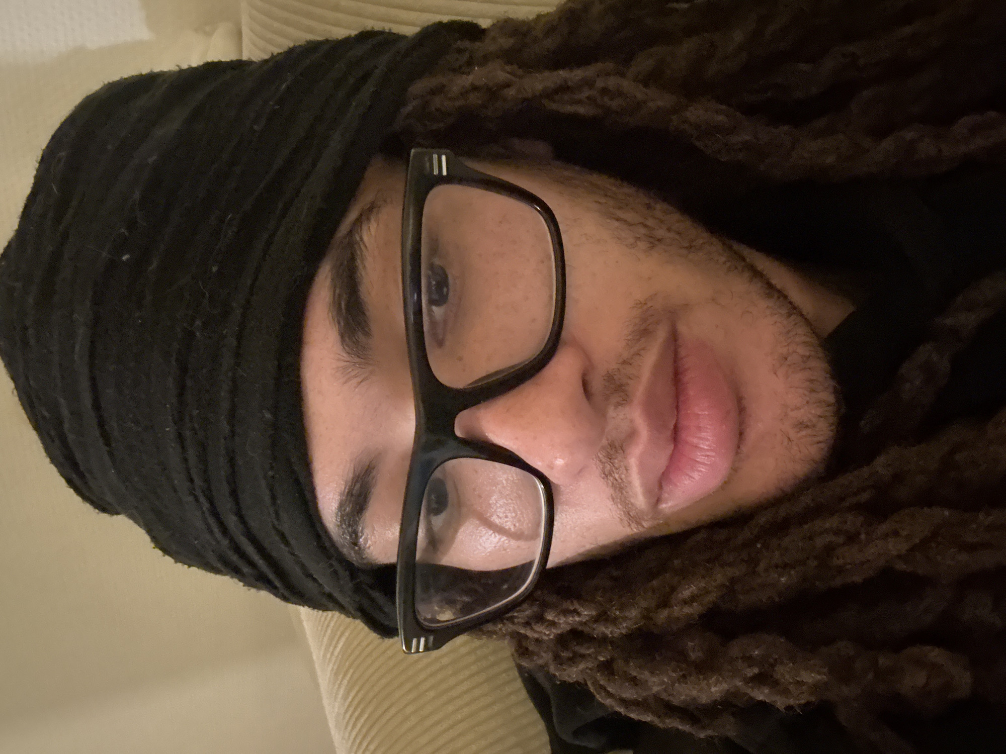 A close-up, indoor portrait shows a person with brown dreadlocks and a black cloth wrapped around their head. They are wearing black-framed glasses and have light skin. The background appears to be a neutral-toned couch. The location cannot be determined.