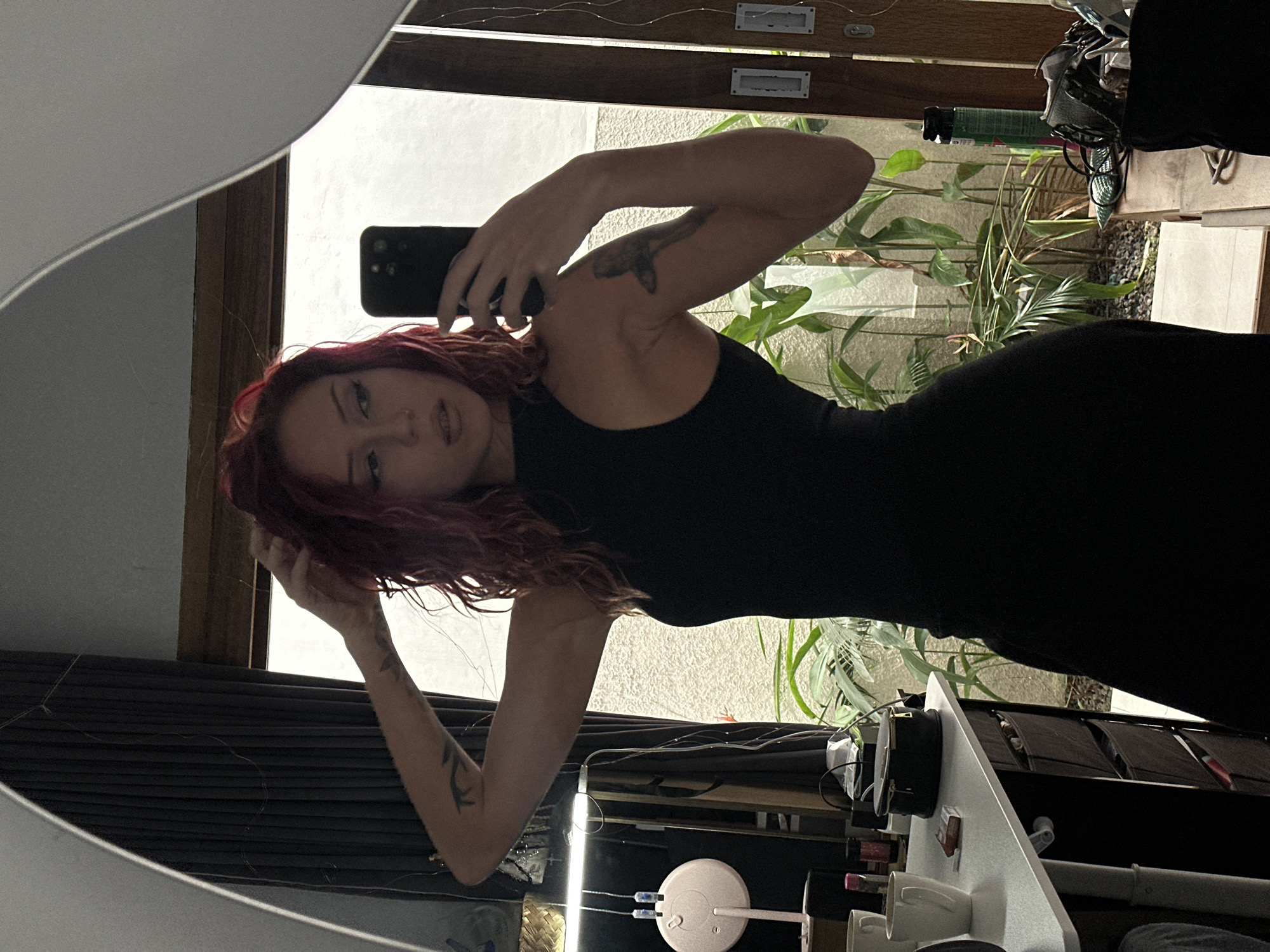 A woman with red hair is taking a selfie in a mirror. She is wearing a black dress. She has tattoos on both arms. Behind her, through a window, are plants and a white wall. A makeup table and other objects are visible in the foreground. The location is North Kuta, Indonesia, as indicated by metadata.
