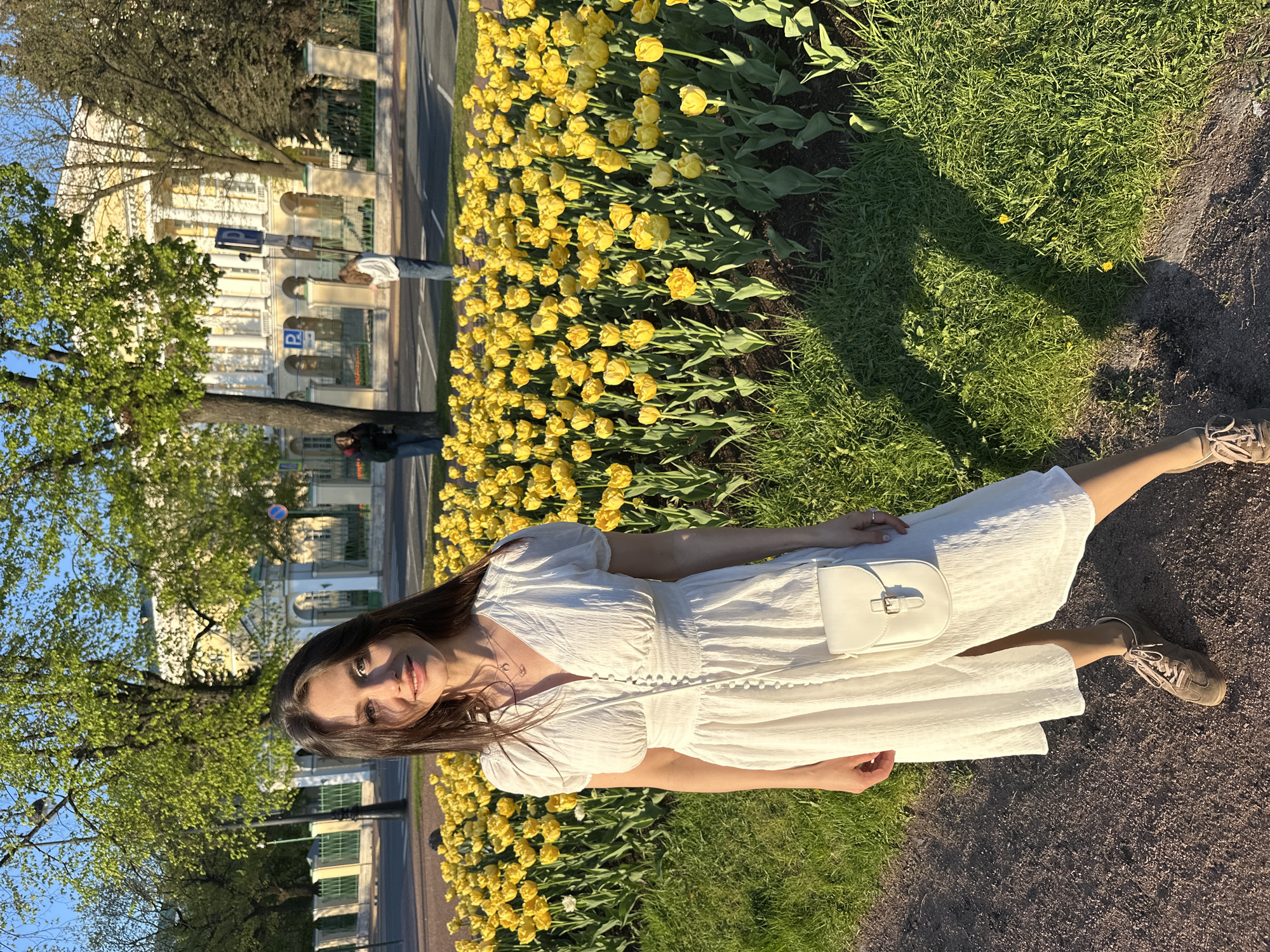 A woman in a white dress lies on a grassy area adjacent to a flower bed of yellow tulips. A narrow strip of asphalt separates the flowers from the grass. Behind the flower bed is a street with a building visible in the background. The scene is outdoors, and the presence of trees and sky suggests it is likely a park area within the city of Saint Petersburg, Russia.
