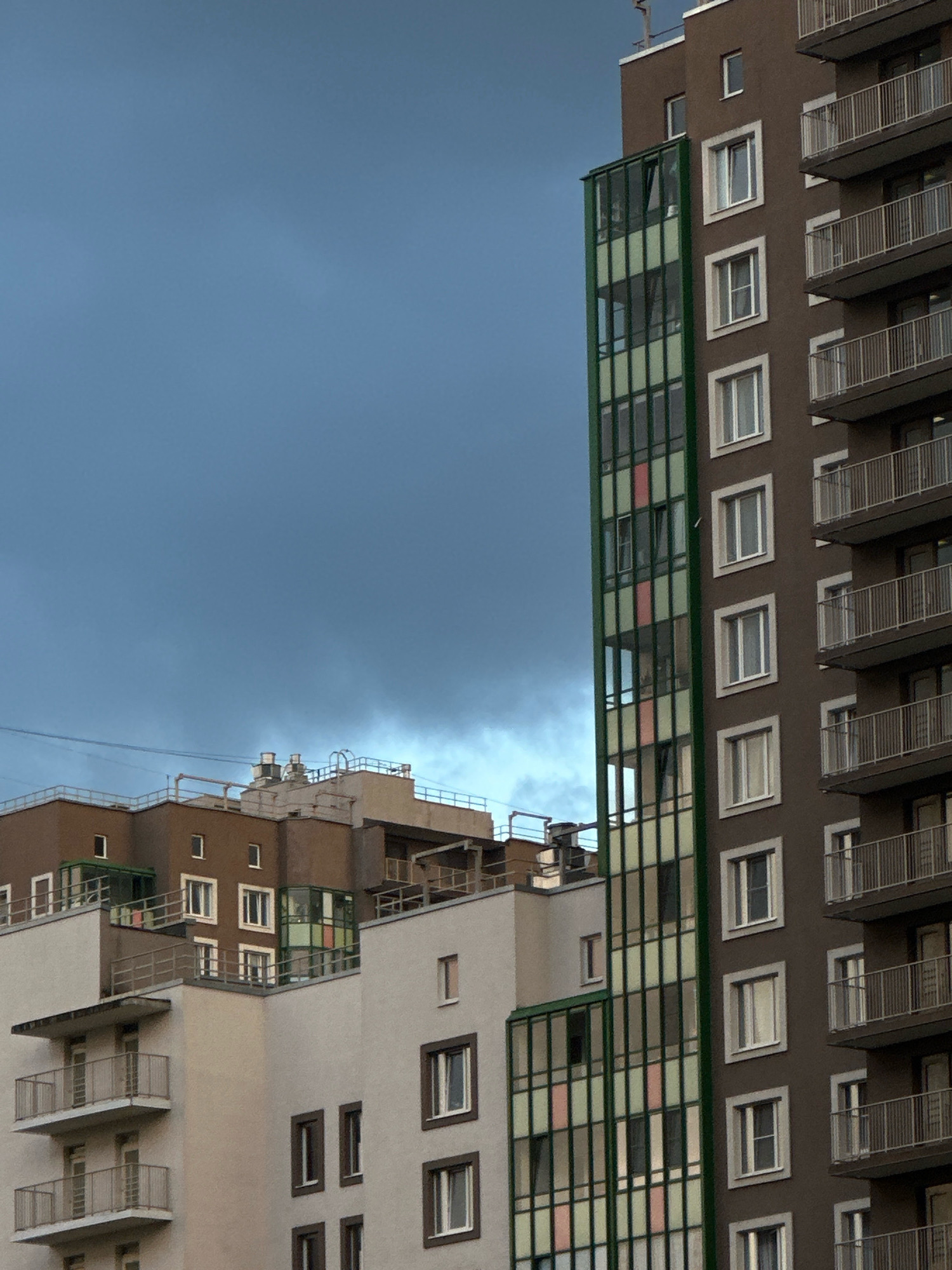 The image captures a close-up view of modern apartment buildings against a cloudy, grey sky. The buildings feature varied architectural styles, with a prominent dark brown high-rise on the right side adorned with multiple balconies and white-framed windows. To the left, a cluster of mid-rise buildings displays a mix of beige and brown facades, some with contrasting green-framed glass enclosures. The scene is devoid of people, focusing solely on the urban architecture and the atmospheric sky.